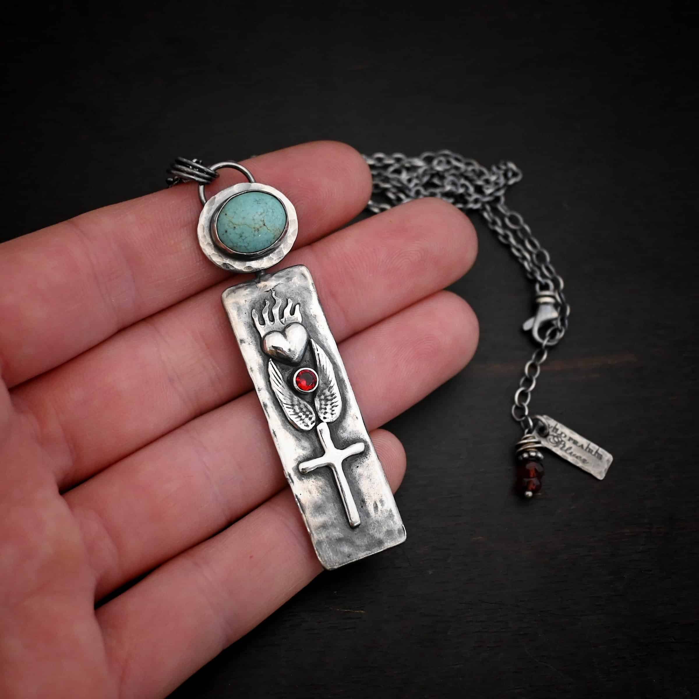 Photo of the "Sacred Heart" Necklace. Necklace is made out of silver, garnet, and turquoise. The pendant is a rectangle shaped silver tag that hangs vertically. It is large. On the top of the rectangle is a silver heart with flames dancing across the top of it. Beneath the heart on the bottom tip of it is a circular small garnet. Beneath this are two silver angel wings, one on each side of the garnet surrounding it. Beneath the angel wings in the center of them and on the bottom of the rectangle pendant is a silver cross soldered to the pendant. There is an oval shaped turquoise soldered to the top of the rectangular pendant. The silver around the turquoise is hammered and oxidized. There is a loop on the back of the turquoise with three jump rings going through it. The chain of the necklace goes through the jump rings. Necklace is lying against the fingertips of a hand.