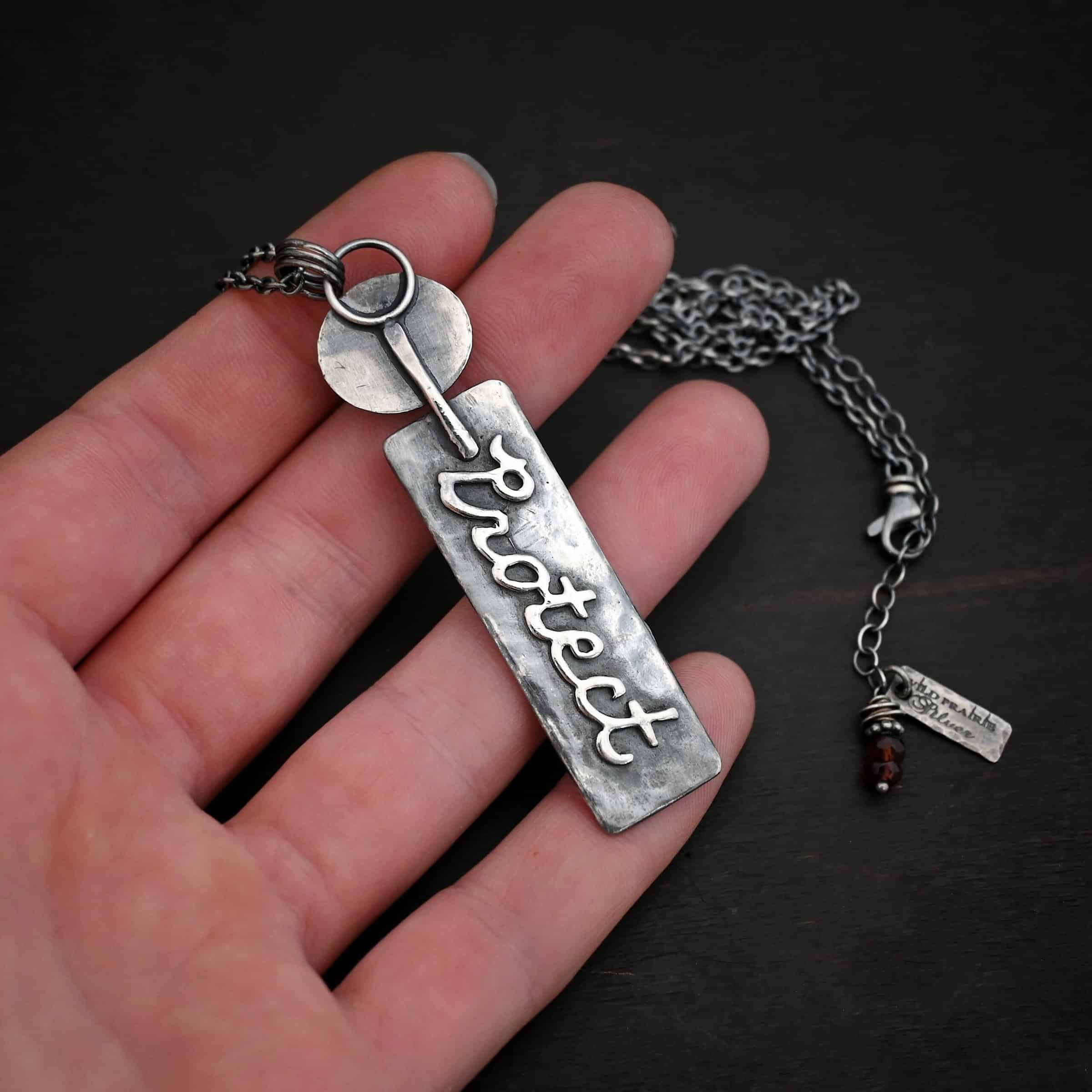 Photo of the back of the "Sacred Heart" Necklace. The top of the pendant starts with an oval shaped piece of silver with a small, but thick stick of silver soldered to the back and going past the bottom. The bottom part of the stick is soldered to the top half of a large silver rectangle. This has the word "Protect" written in cursive and soldered to the back of it. Necklace is lying against the fingertips of a hand.