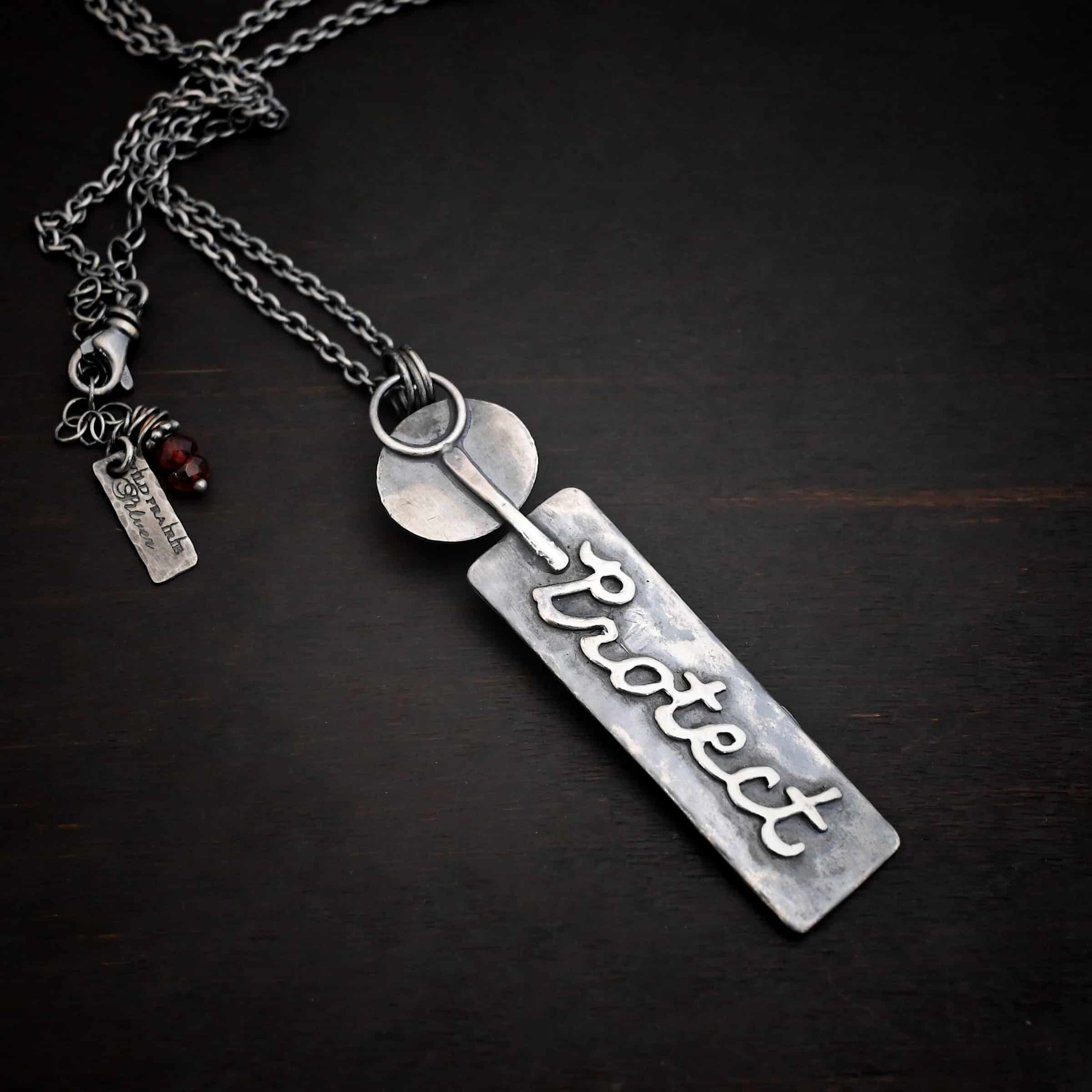Photo of the back of the "Sacred Heart" Necklace. The top of the pendant starts with an oval shaped piece of silver with a small, but thick stick of silver soldered to the back and going past the bottom. The bottom part of the stick is soldered to the top half of a large silver rectangle. This has the word "Protect" written in cursive and soldered to the back of it. Necklace is lying against a dark background.