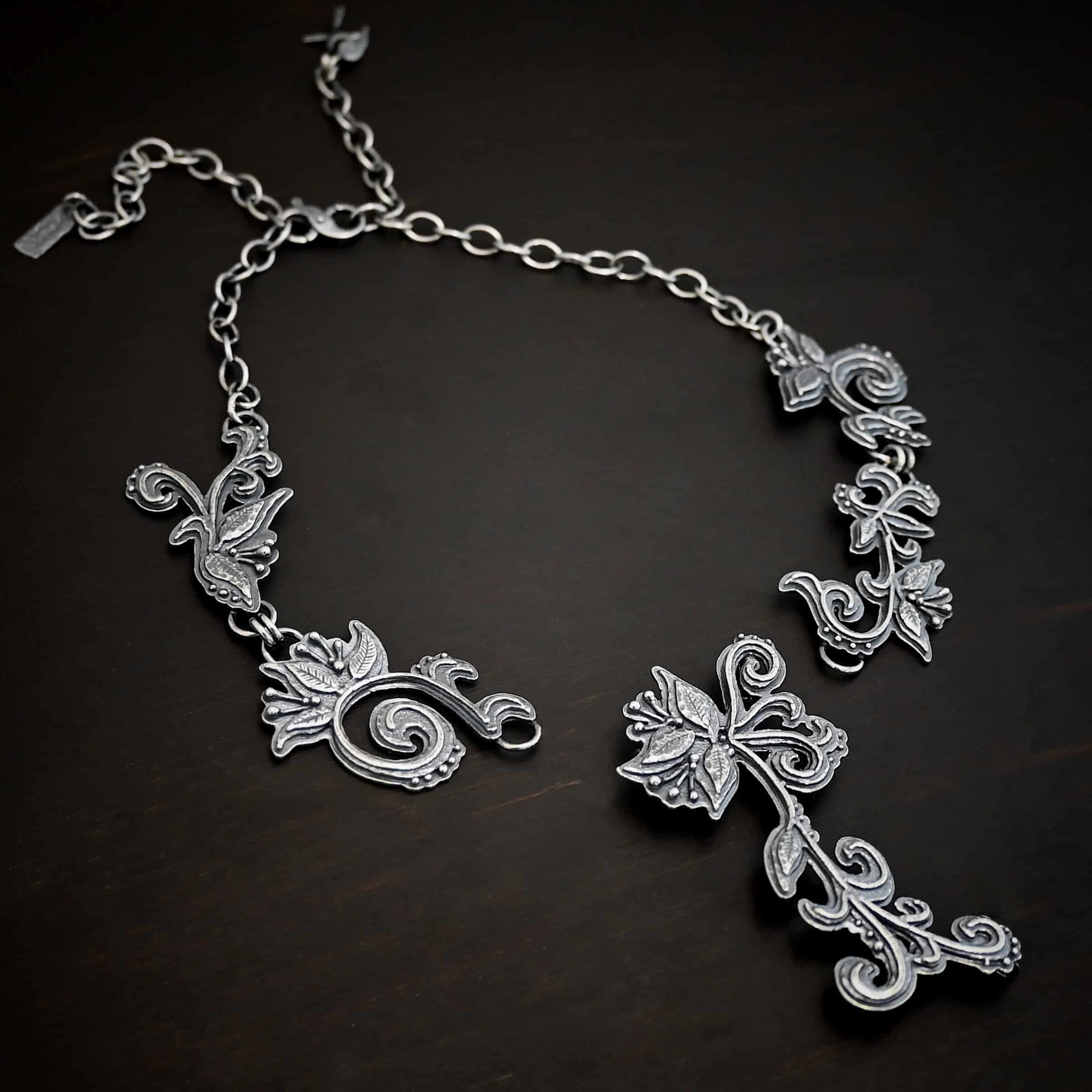 Paisley Necklace Photo of the "Paisley" Necklace. Made out of sterling silver. It is a large collar necklace. The entire necklace is made out of pendants that have sterling silver leaves and silver ball detail around the swirling pendants. The bottom of the necklace has a long, vine like pieces of silver that hangs down, with silver leaves at the top and in the center, and small swirls on the bottom. Necklace is lying against a dark background.