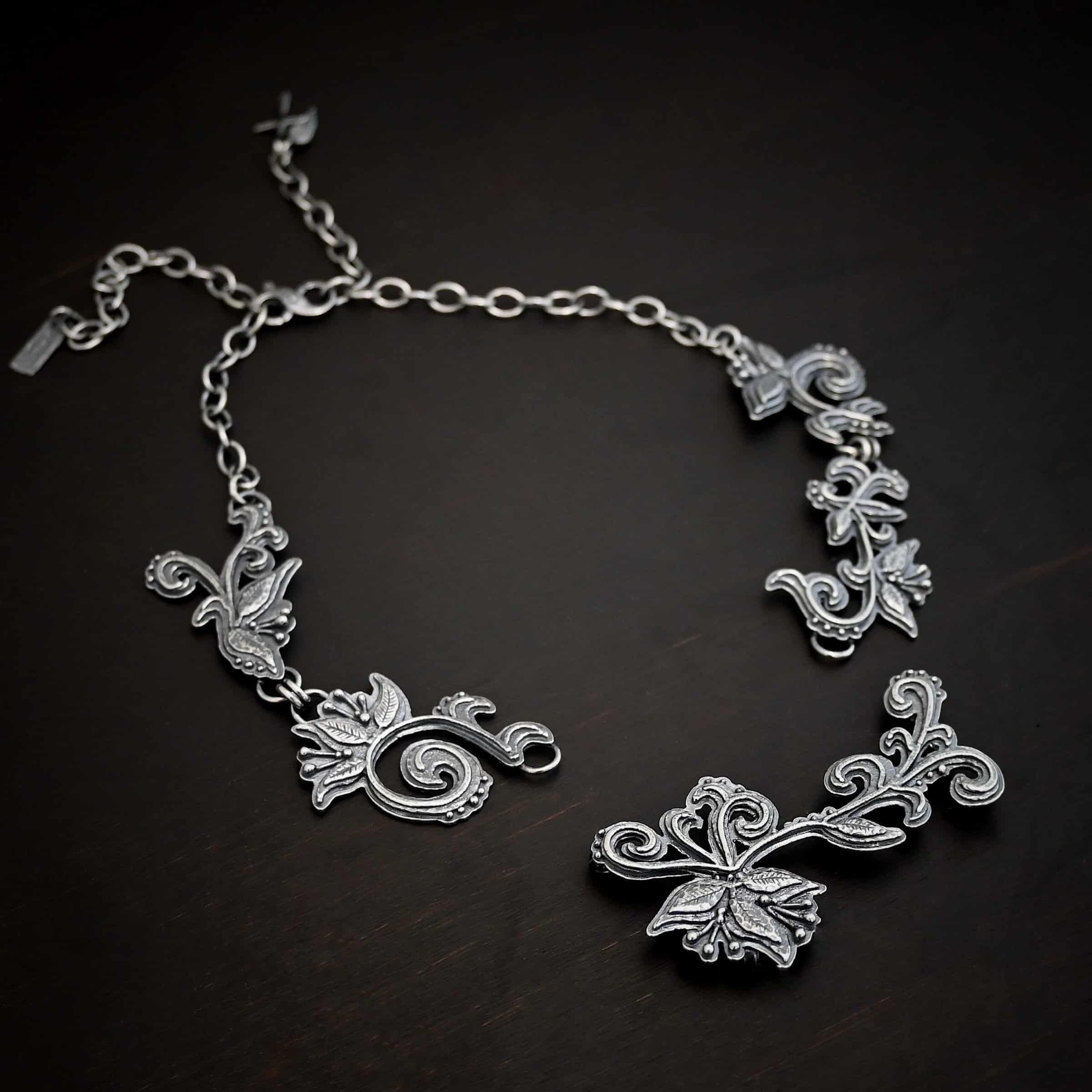 Paisley Necklace Photo of the "Paisley" Necklace. Made out of sterling silver. It is a large collar necklace. The entire necklace is made out of pendants that have sterling silver leaves and silver ball detail around the swirling pendants. The bottom of the necklace has a long, vine like pieces of silver that hangs down, with silver leaves at the top and in the center, and small swirls on the bottom. Necklace is lying against a dark background.
