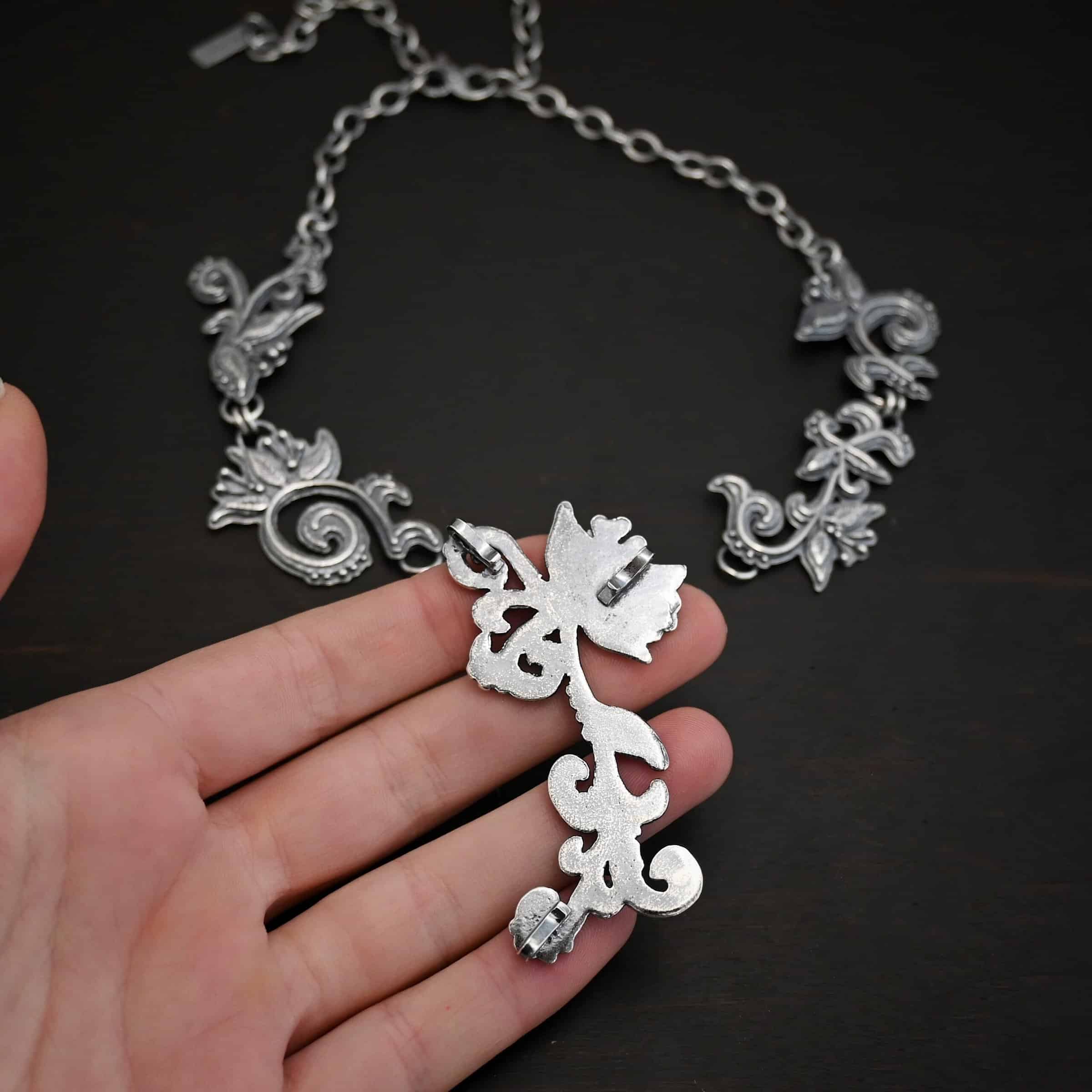 Paisley Necklace Photo of the "Paisley" Necklace. Made out of sterling silver. It is a large collar necklace. The entire necklace is made out of pendants that have sterling silver leaves and silver ball detail around the swirling pendants. The bottom of the necklace has a long, vine like pieces of silver that hangs down, with silver leaves at the top and in the center, and small swirls on the bottom. Necklace is lying against the fingertips of a hand.