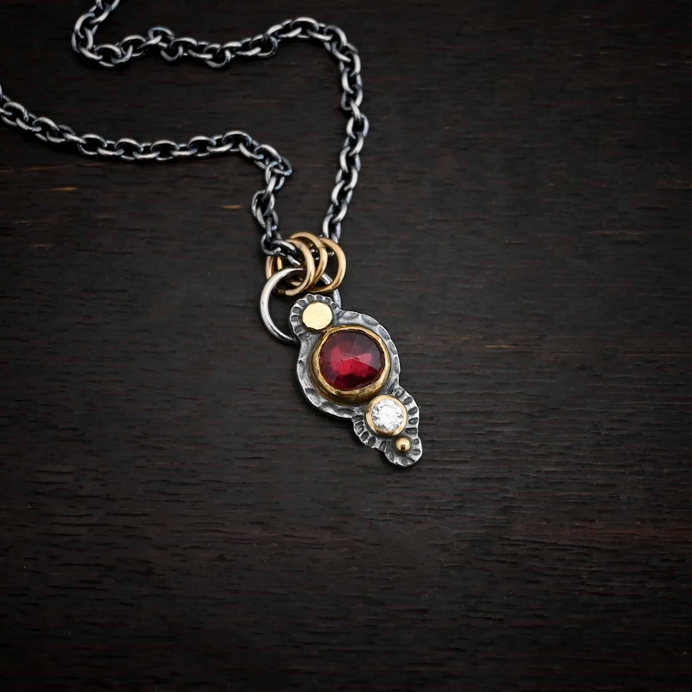 Photo of the "Champagne Blush" Necklace. There is a gold dot at the top, then a small circular garnet set in gold, and then a small diamond that is set in gold, with a gold ball on the very bottom of the pendant. There are three gold jump rings attached to a loop on the top. The chain goes through the jump rings. Necklace is lying against a dark background.
