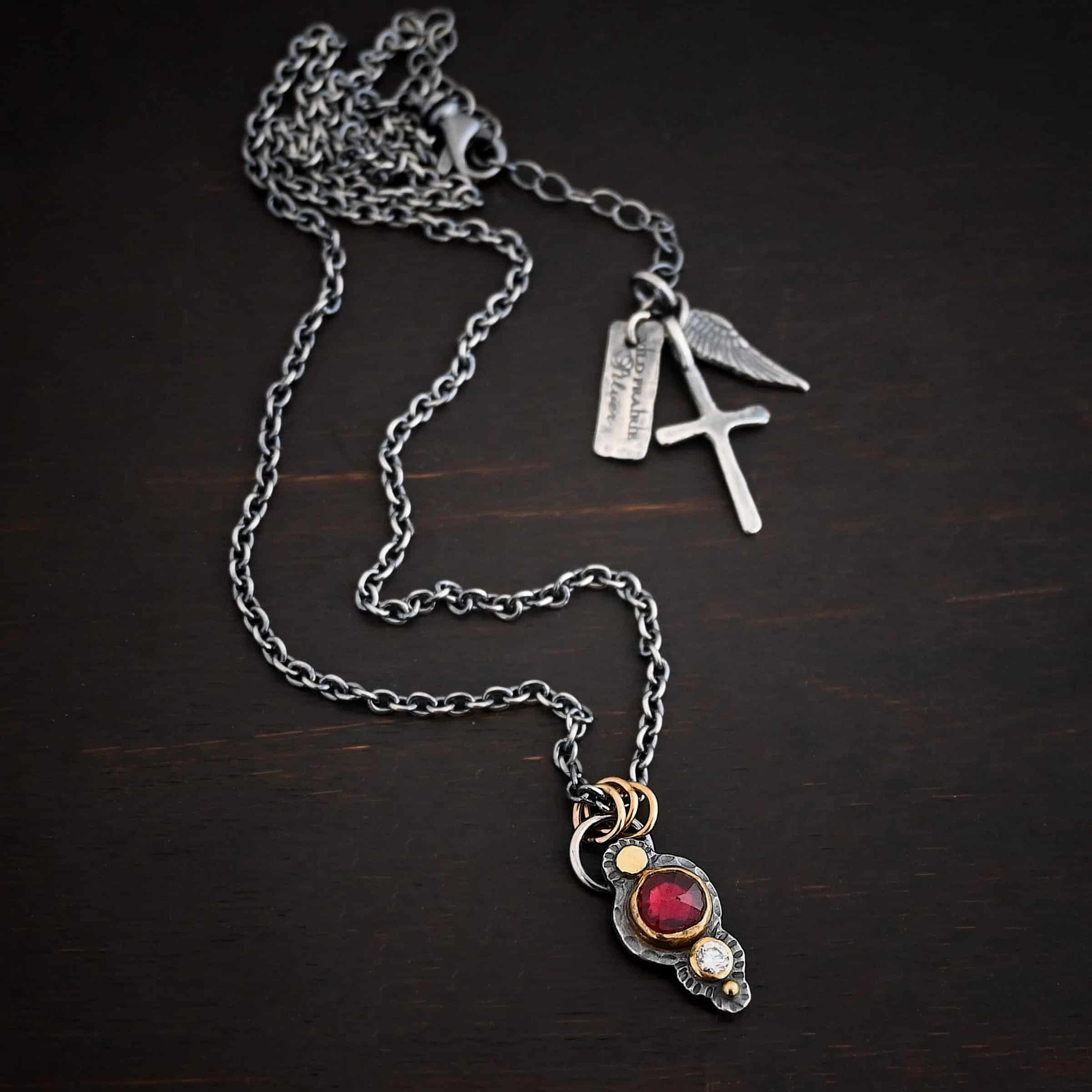 Photo of the "Champagne Blush" Necklace. There is a gold dot at the top, then a small circular garnet set in gold, and then a small diamond that is set in gold, with a gold ball on the very bottom of the pendant. There are three gold jump rings attached to a loop on the top. The chain goes through the jump rings. Necklace is lying against a dark background.