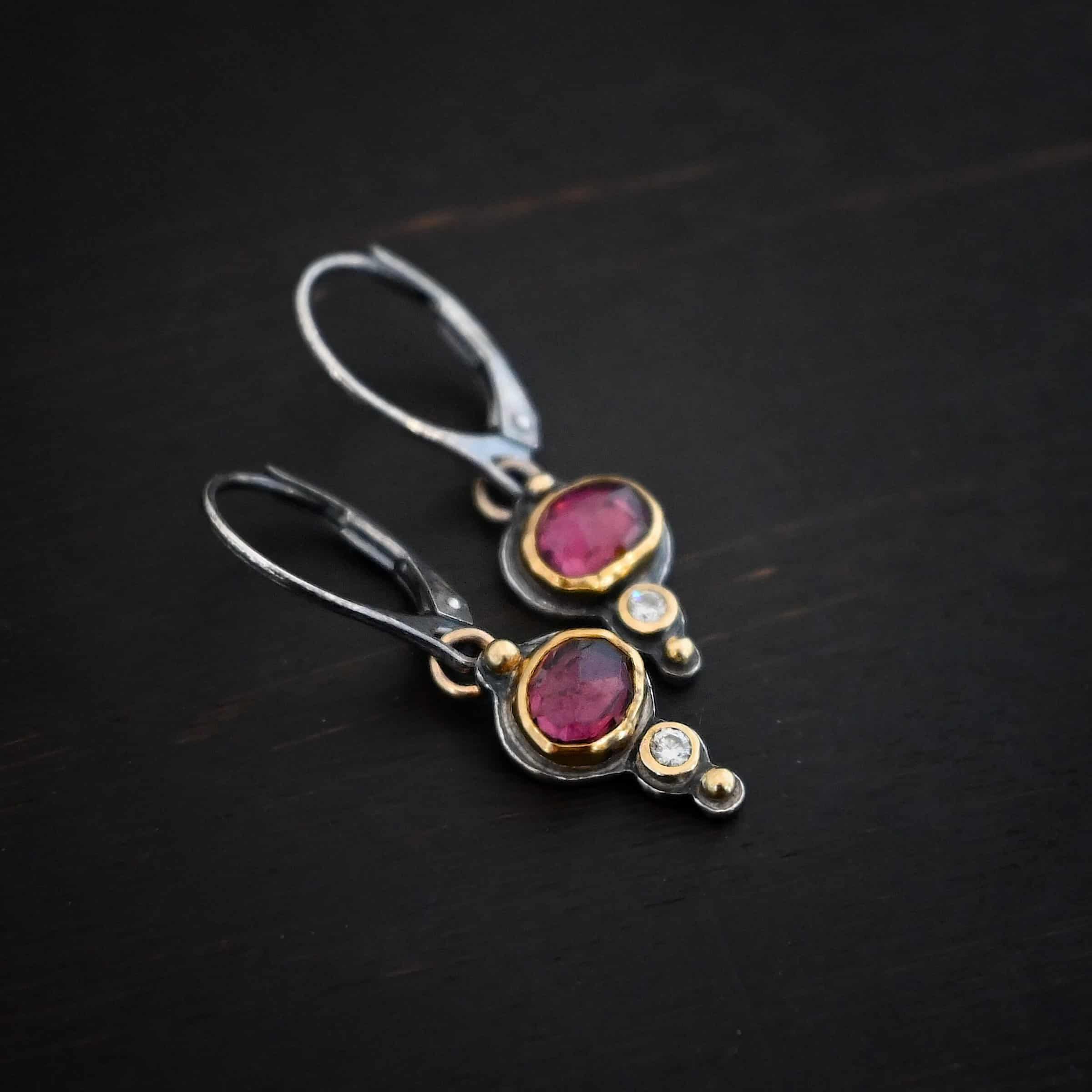 Champagne Blush Earrings Photo of the "Champagne Blush" Earrings. Each earring has a ball of gold on the top, then an oval garnet, then a small diamond, and finally another ball of gold. The garnets and diamonds are set in gold. Earrings have sterling silver leverback ear wires. Earrings are lying against a dark background.