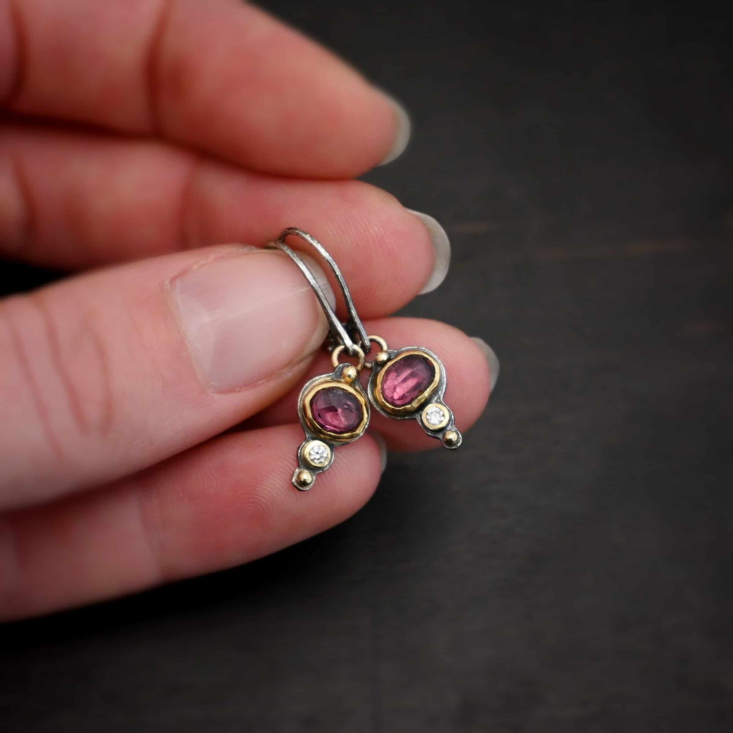 Champagne Blush Earrings Photo of the "Champagne Blush" Earrings. Each earring has a ball of gold on the top, then an oval garnet, then a small diamond, and finally another ball of gold. The garnets and diamonds are set in gold. Earrings have sterling silver leverback ear wires and are held in the fingertips of a hand.