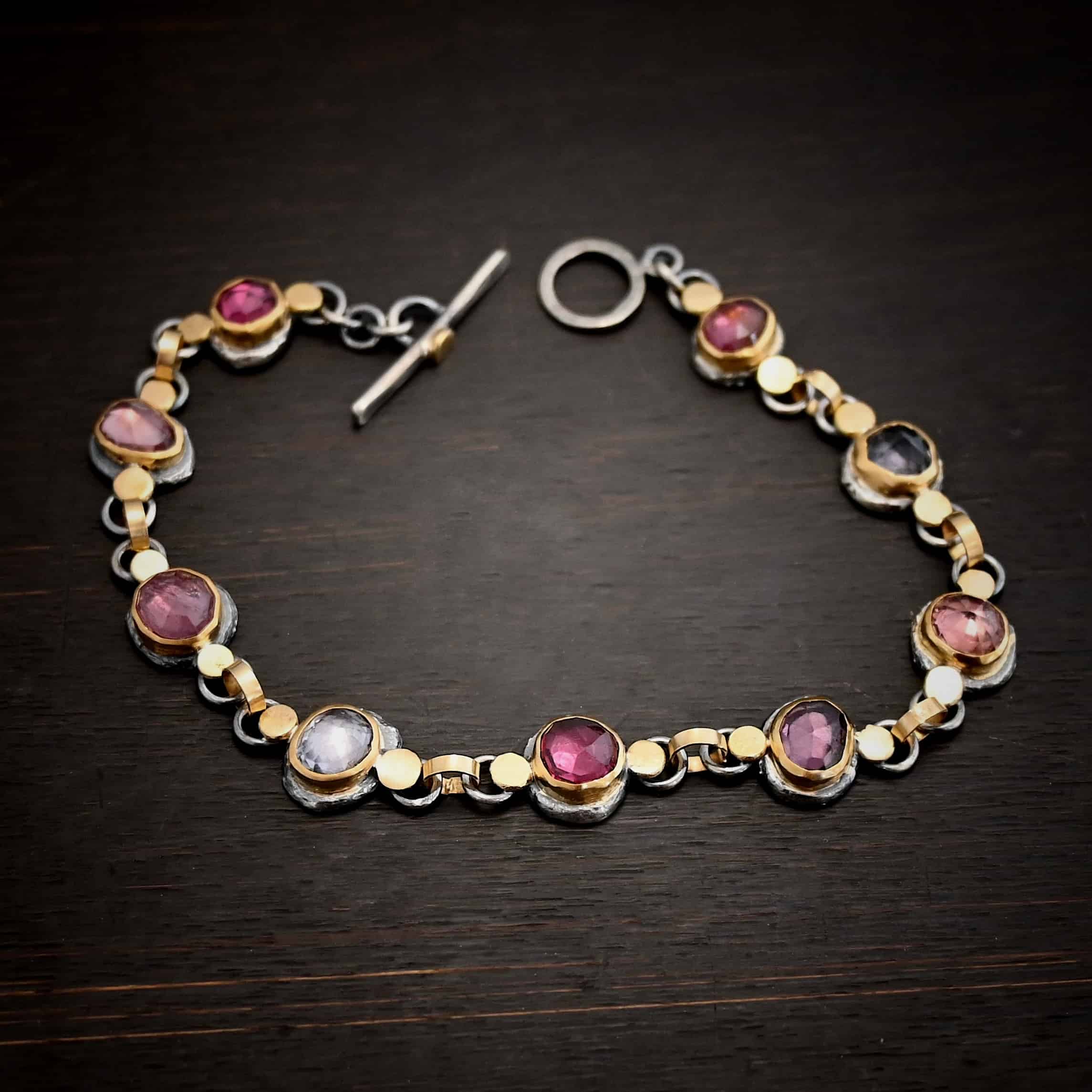 Photo of the "Sable" Bracelet. Made out of sterling silver & 18k gold & sapphires. There are nine sapphires, each a pink, clear, or purple color. Each one is set in gold, and has a large gold dot on each side of it, making for 18 gold dots. Each sapphire has two sterling silver loops soldered to the back of it, and each loop has a gold jump ring to connect all of the sapphires to each other. Bracelet has a toggle clasp and is lying against a dark background.