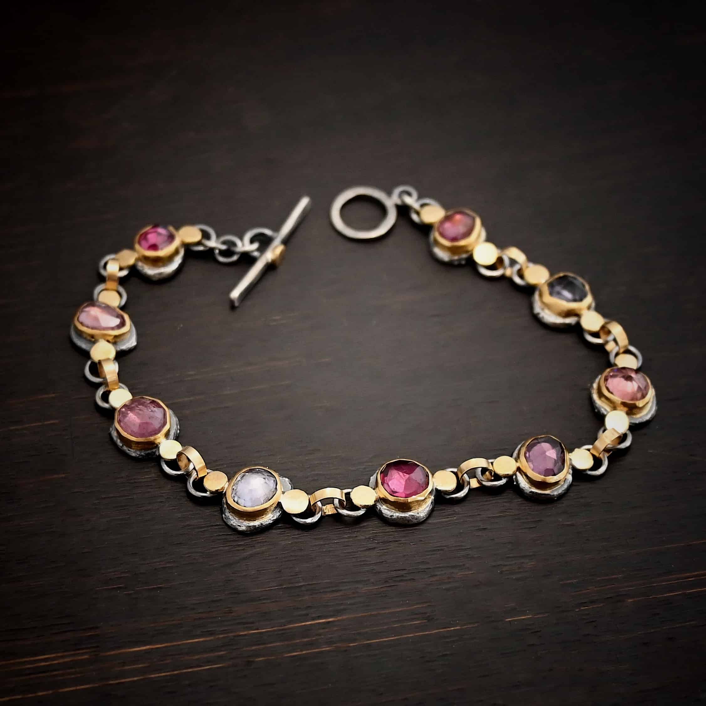 Photo of the "Sable" Bracelet. Made out of sterling silver & 18k gold & sapphires. There are nine sapphires, each a pink, clear, or purple color. Each one is set in gold, and has a large gold dot on each side of it, making for 18 gold dots. Each sapphire has two sterling silver loops soldered to the back of it, and each loop has a gold jump ring to connect all of the sapphires to each other. Bracelet has a toggle clasp and is lying against a dark background.