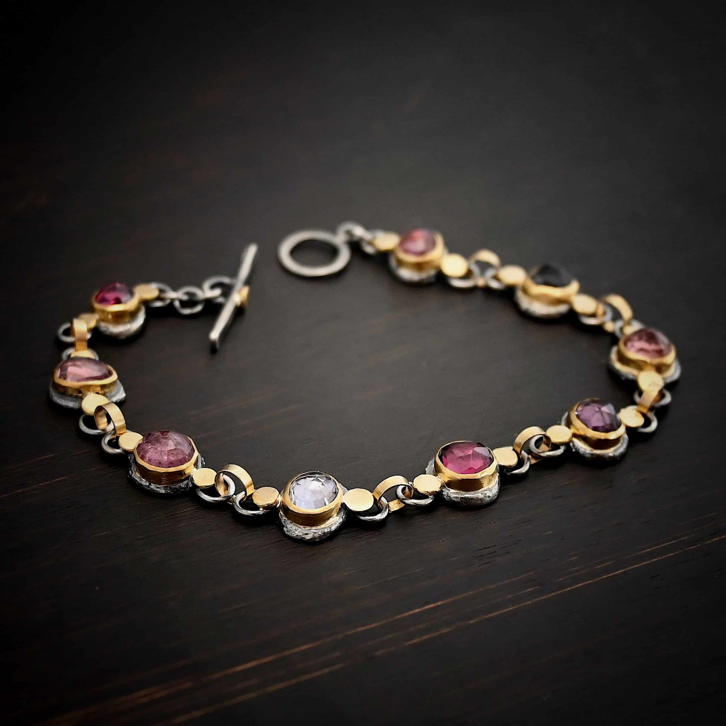 Photo of the "Sable" Bracelet. Made out of sterling silver & 18k gold & sapphires. There are nine sapphires, each a pink, clear, or purple color. Each one is set in gold, and has a large gold dot on each side of it, making for 18 gold dots. Each sapphire has two sterling silver loops soldered to the back of it, and each loop has a gold jump ring to connect all of the sapphires to each other. Bracelet has a toggle clasp and is lying against a dark background.