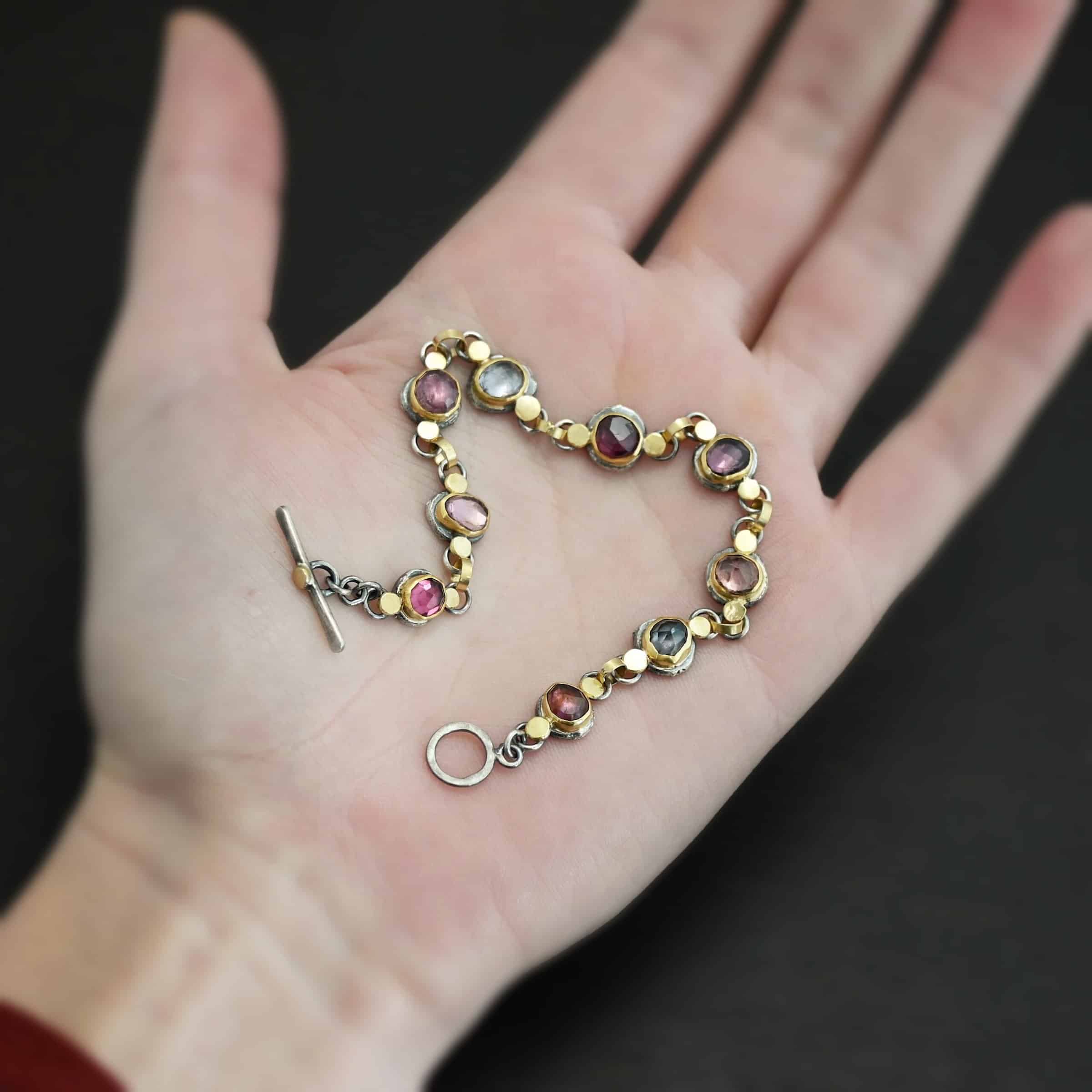 Photo of the "Sable" Bracelet. Made out of sterling silver & 18k gold & sapphires. There are nine sapphires, each a pink, clear, or purple color. Each one is set in gold, and has a large gold dot on each side of it, making for 18 gold dots. Each sapphire has two sterling silver loops soldered to the back of it, and each loop has a gold jump ring to connect all of the sapphires to each other. Bracelet has a toggle clasp and is lying against the palm of a hand.