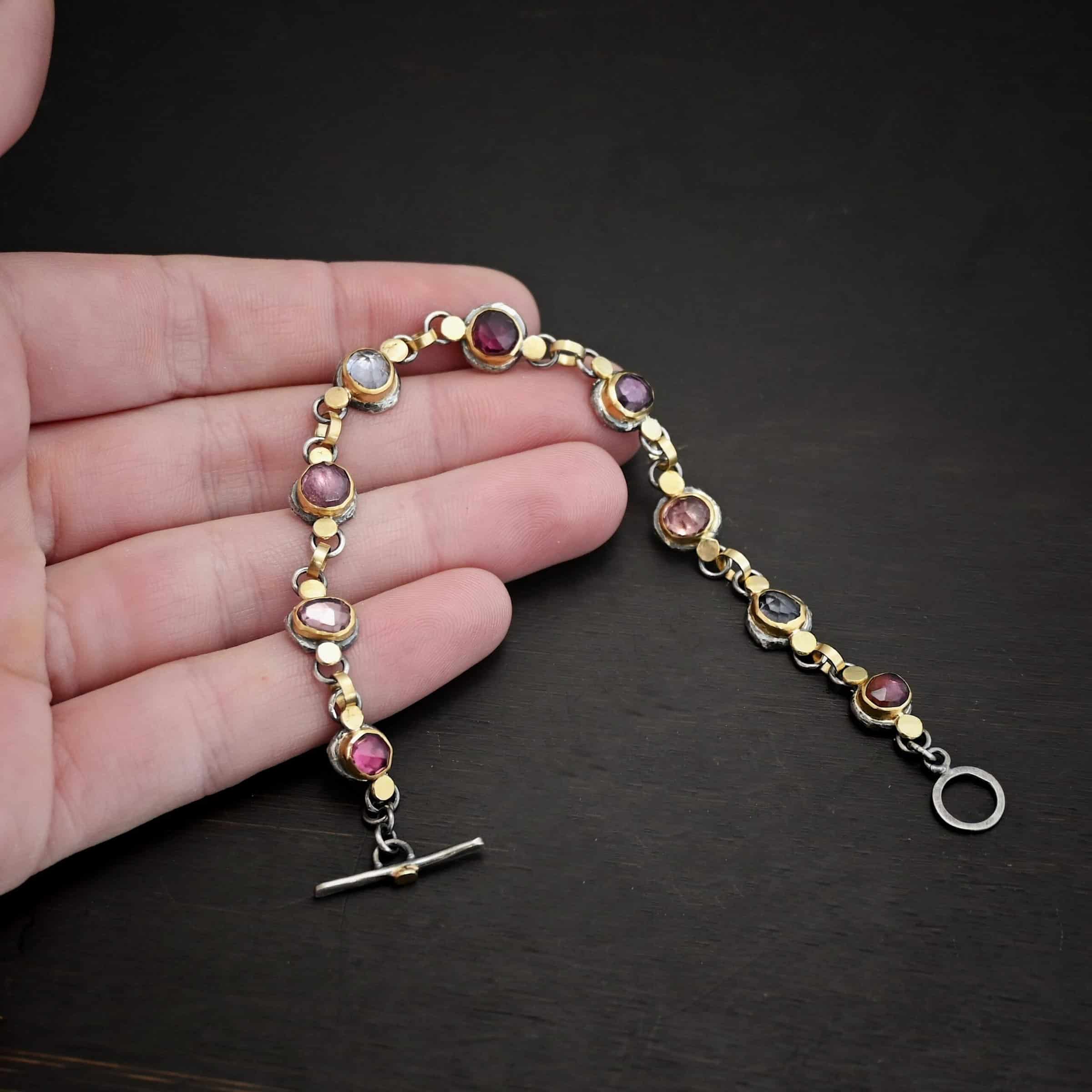 Photo of the "Sable" Bracelet. Made out of sterling silver & 18k gold & sapphires. There are nine sapphires, each a pink, clear, or purple color. Each one is set in gold, and has a large gold dot on each side of it, making for 18 gold dots. Each sapphire has two sterling silver loops soldered to the back of it, and each loop has a gold jump ring to connect all of the sapphires to each other. Bracelet has a toggle clasp and is lying against the fingertips of a hand.