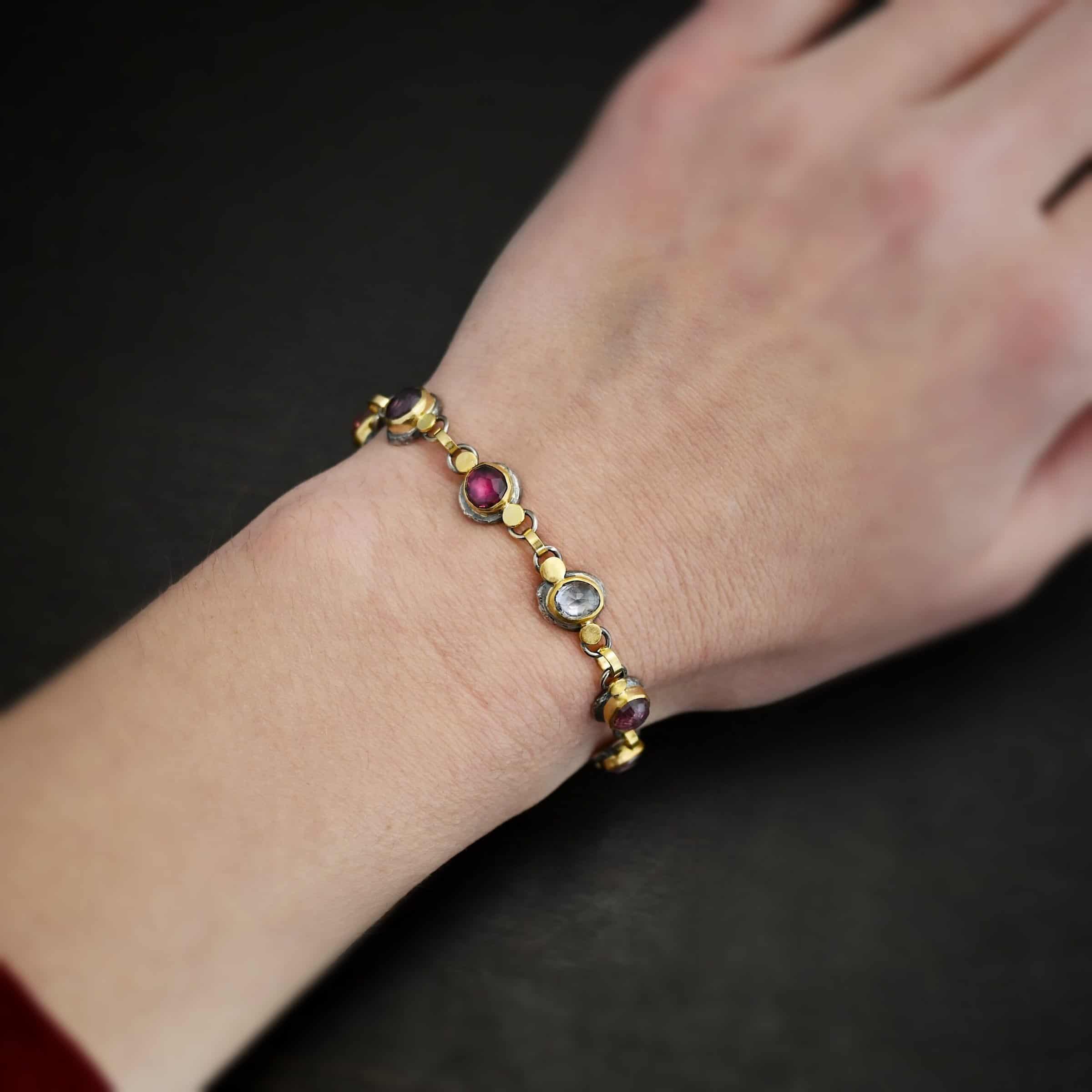 Photo of the "Sable" Bracelet. Made out of sterling silver & 18k gold & sapphires. There are nine sapphires, each a pink, clear, or purple color. Each one is set in gold, and has a large gold dot on each side of it, making for 18 gold dots. Each sapphire has two sterling silver loops soldered to the back of it, and each loop has a gold jump ring to connect all of the sapphires to each other. Bracelet has a toggle clasp and is worn on the wrist by a model.