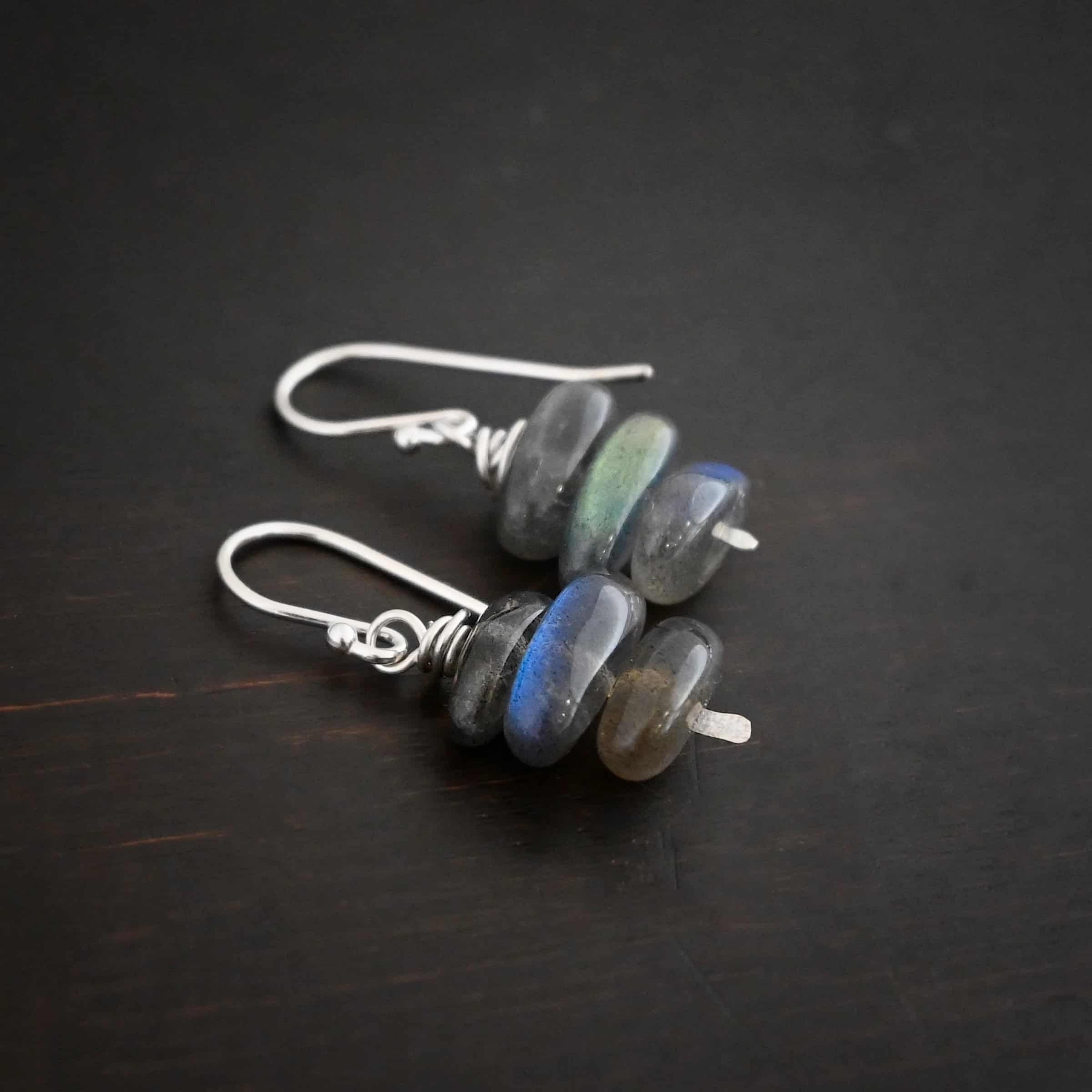 Indigo Moon Earrings Photo of the "Indigo Moon" Earrings. Made out of sterling silver and labradorite. Earrings have french hook ear wires. Attached to the bottom of the ear wires is a small stick of sterling silver with a loop on the top and silver that swirls around the bottom of the loop three times and a hammered paddle on the bottom. In the center of the paddle and the swirling silver are three long oval pieces of labradorite that are horizontal. Earrings are lying against a dark background.