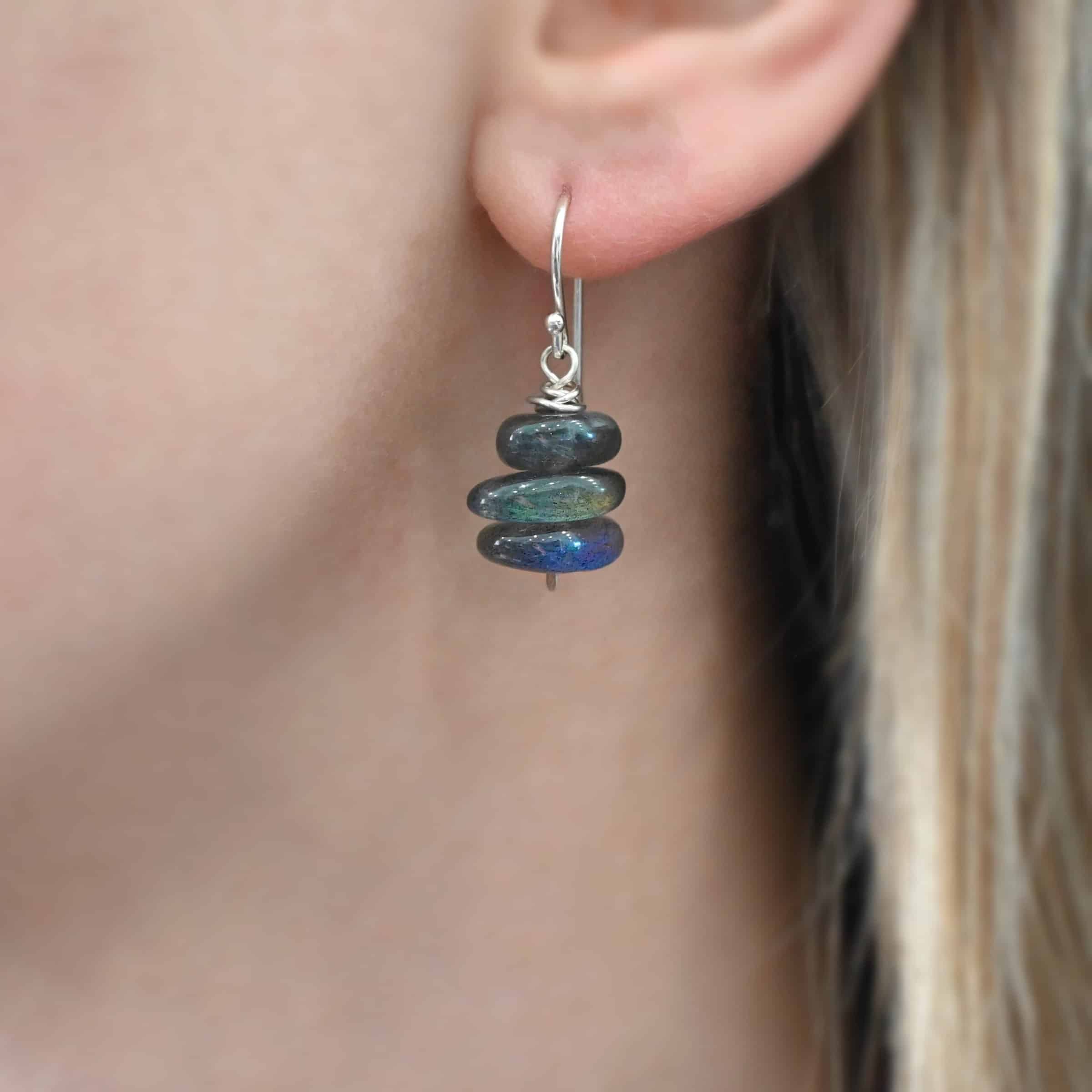 Indigo Moon Earrings Photo of the "Indigo Moon" Earring as shown on a model. Made out of sterling silver and labradorite. Earring has a french hook ear wire. Attached to the bottom of the ear wires is a small stick of sterling silver with a loop on the top and silver that swirls around the bottom of the loop three times and a hammered paddle on the bottom. In the center of the paddle and the swirling silver are three long oval pieces of labradorite that are horizontal.
