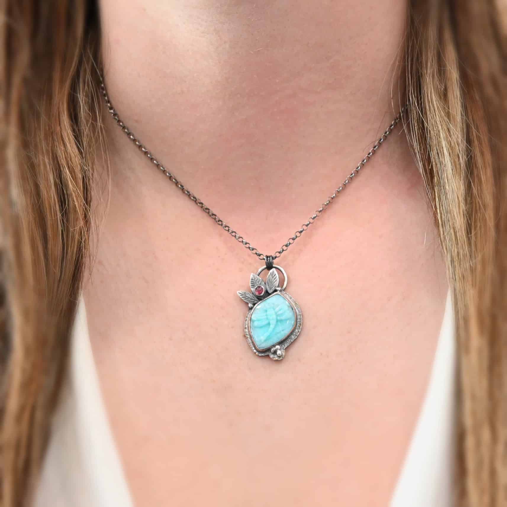 Close up of the "Libelle" Necklace. Handmade out of sterling silver, amazonite, and pink tourmaline. The amazonite is the main stone, and it has a dragonfly carved into it. The pink tourmaline is near the top of the necklace on the left side. It is set in an engraved sterling silver leaf, which is surrounded by two other sterling silver leaves. There are three small dots on a circle near the bottom to represent a lily pad/ The necklace is hanging on a sterling silver oxidized chain. Chain is set at 16" long on the model. Model is wearing a white v-neck shirt.