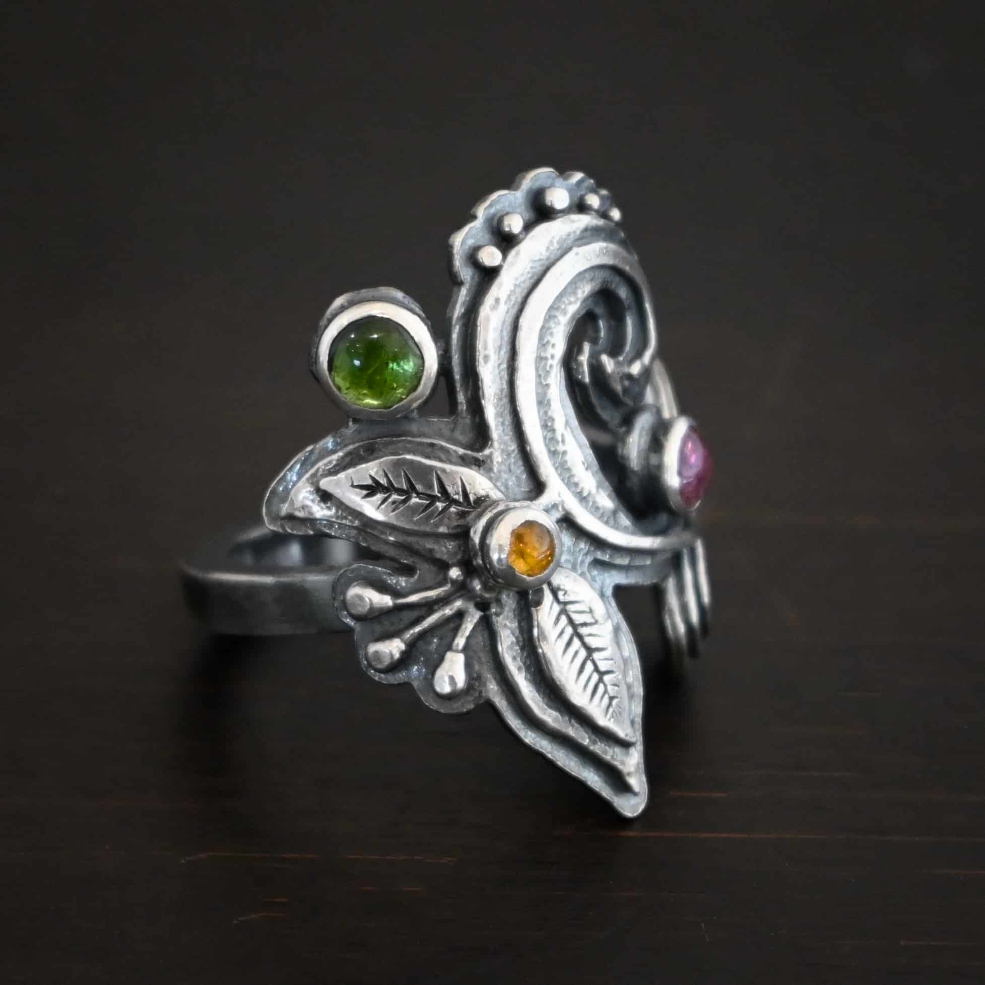 Purple Rain Ring Photo of the "Purple Rain" Ring. Made out of sterling silver and tourmaline. There are three tourmalines, a pink, dark yellow, and green tourmaline. The ring has silver swirls with silver engraved leaves and lines attached to the bottom and top of the swirls. The tourmalines are scattered and there is silver ball detail on the ring as well. Ring is sitting against a dark background.