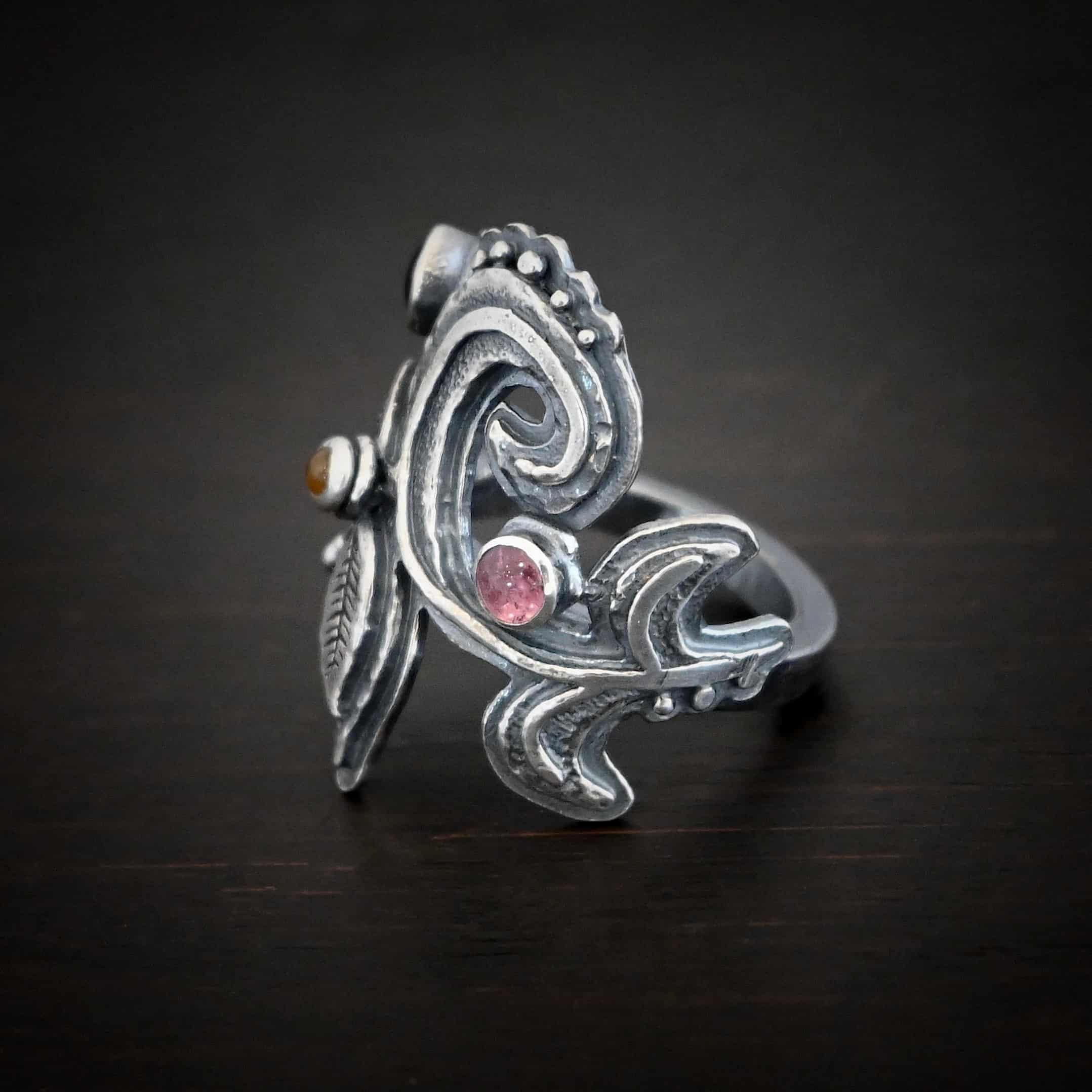 Purple Rain Ring Photo of the "Purple Rain" Ring. Made out of sterling silver and tourmaline. There are three tourmalines, a pink, dark yellow, and green tourmaline. The ring has silver swirls with silver engraved leaves and lines attached to the bottom and top of the swirls. The tourmalines are scattered and there is silver ball detail on the ring as well. Ring is sitting against a dark background.