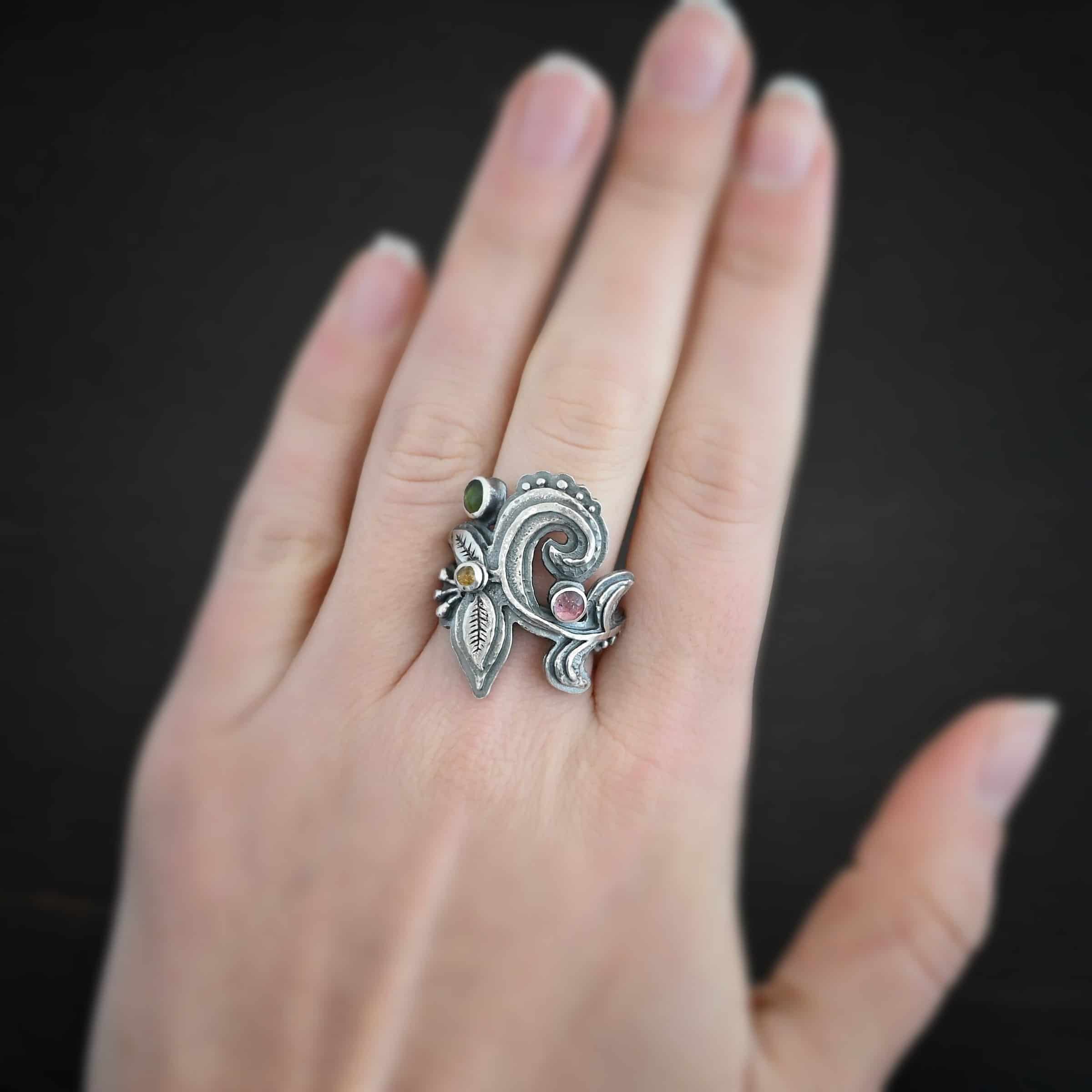 Purple Rain Ring Photo of the "Purple Rain" Ring. Made out of sterling silver and tourmaline. There are three tourmalines, a pink, dark yellow, and green tourmaline. The ring has silver swirls with silver engraved leaves and lines attached to the bottom and top of the swirls. The tourmalines are scattered and there is silver ball detail on the ring as well. Ring is worn on a models middle finger to demonstrate size.