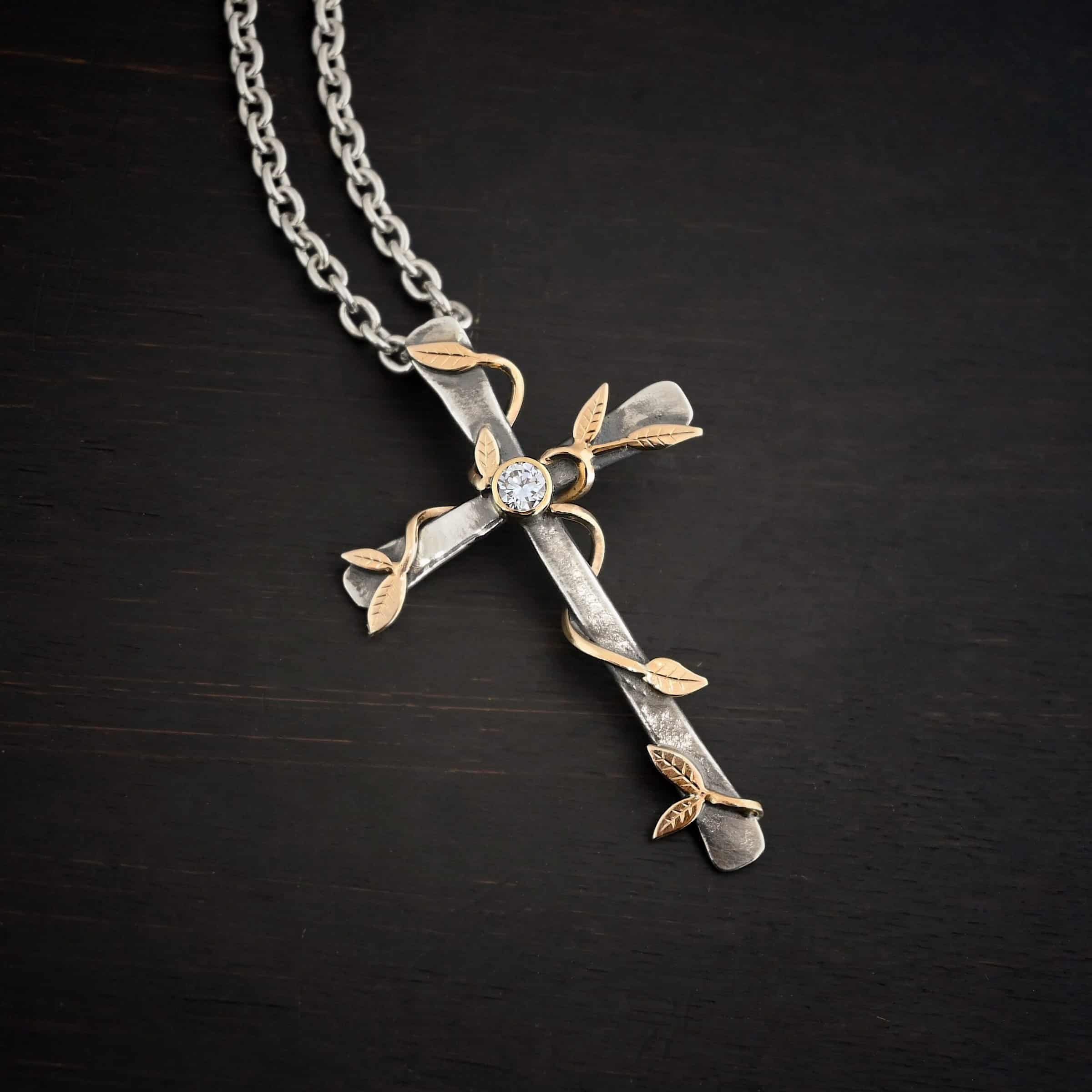 Photo of a custom "He Is Risen" Necklace. There is a large sized, smooth, oxidized, cross pendant. The center has a diamond set in gold. There are gold vines with leaves wrapped around the cross. Necklace is lying against a dark background.