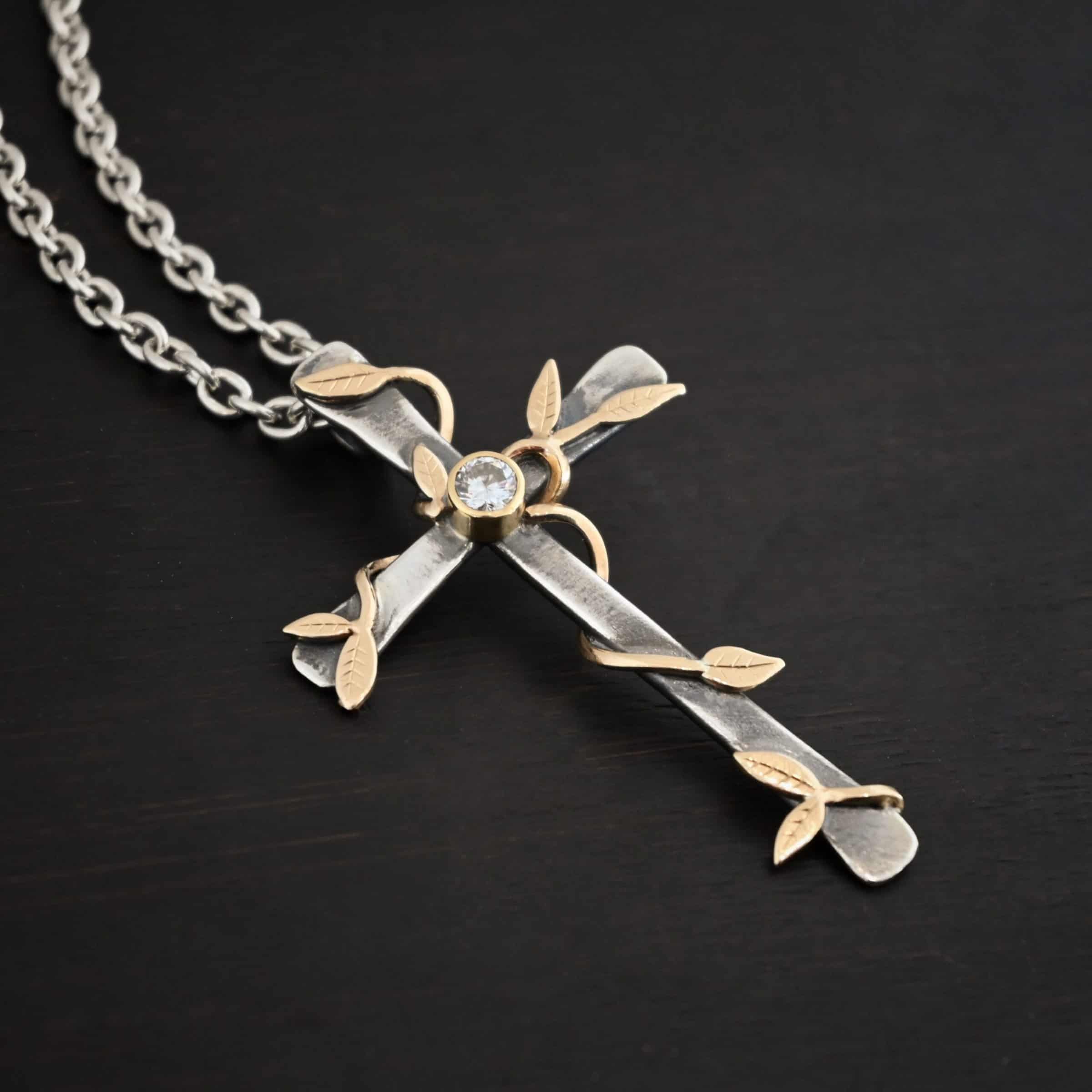 Photo of a custom "He Is Risen" Necklace. There is a large sized, smooth, oxidized, cross pendant. The center has a diamond set in gold. There are gold vines with leaves wrapped around the cross. Necklace is lying against a dark background.