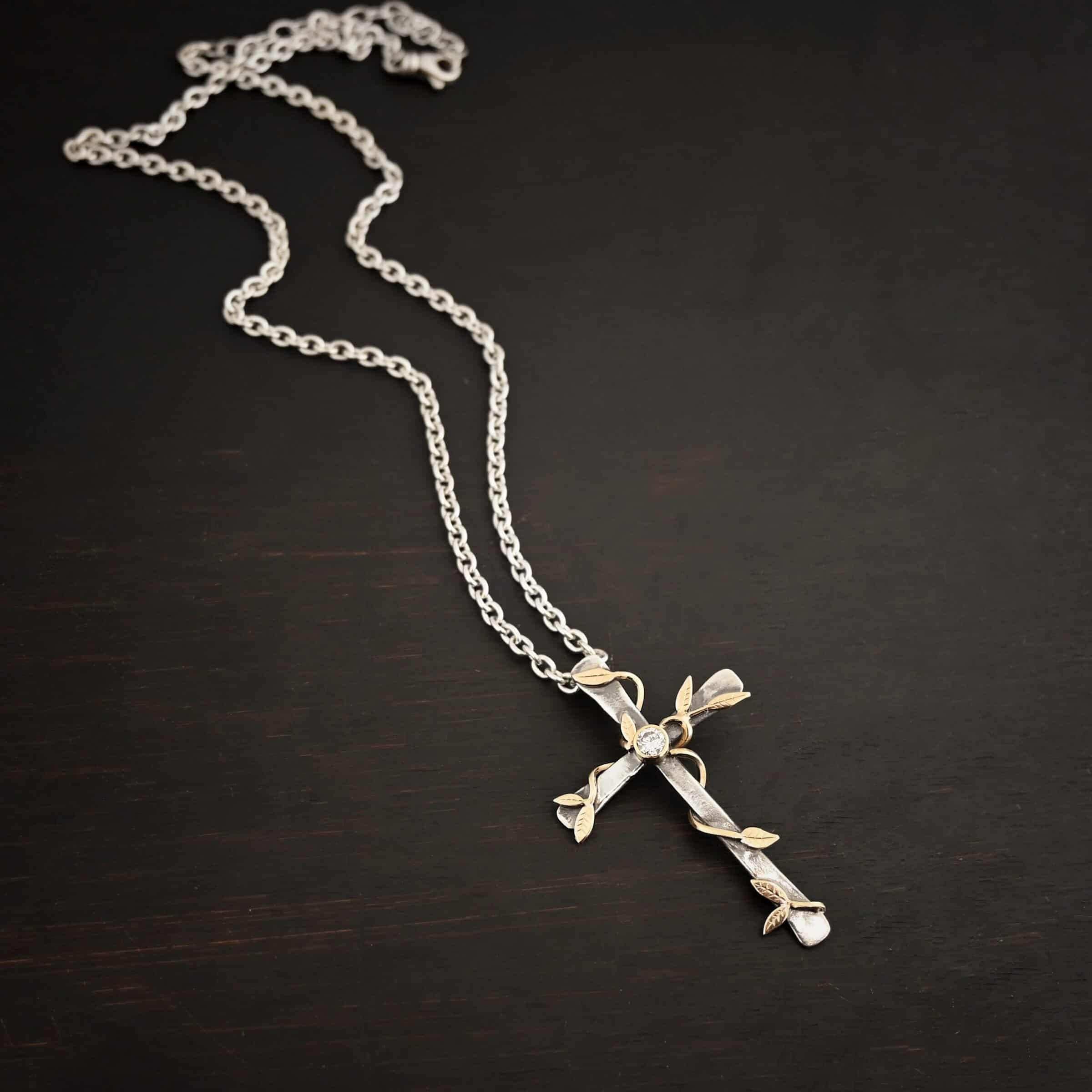 Photo of a custom "He Is Risen" Necklace. There is a large sized, smooth, oxidized, cross pendant. The center has a diamond set in gold. There are gold vines with leaves wrapped around the cross. Necklace is lying against a dark background.