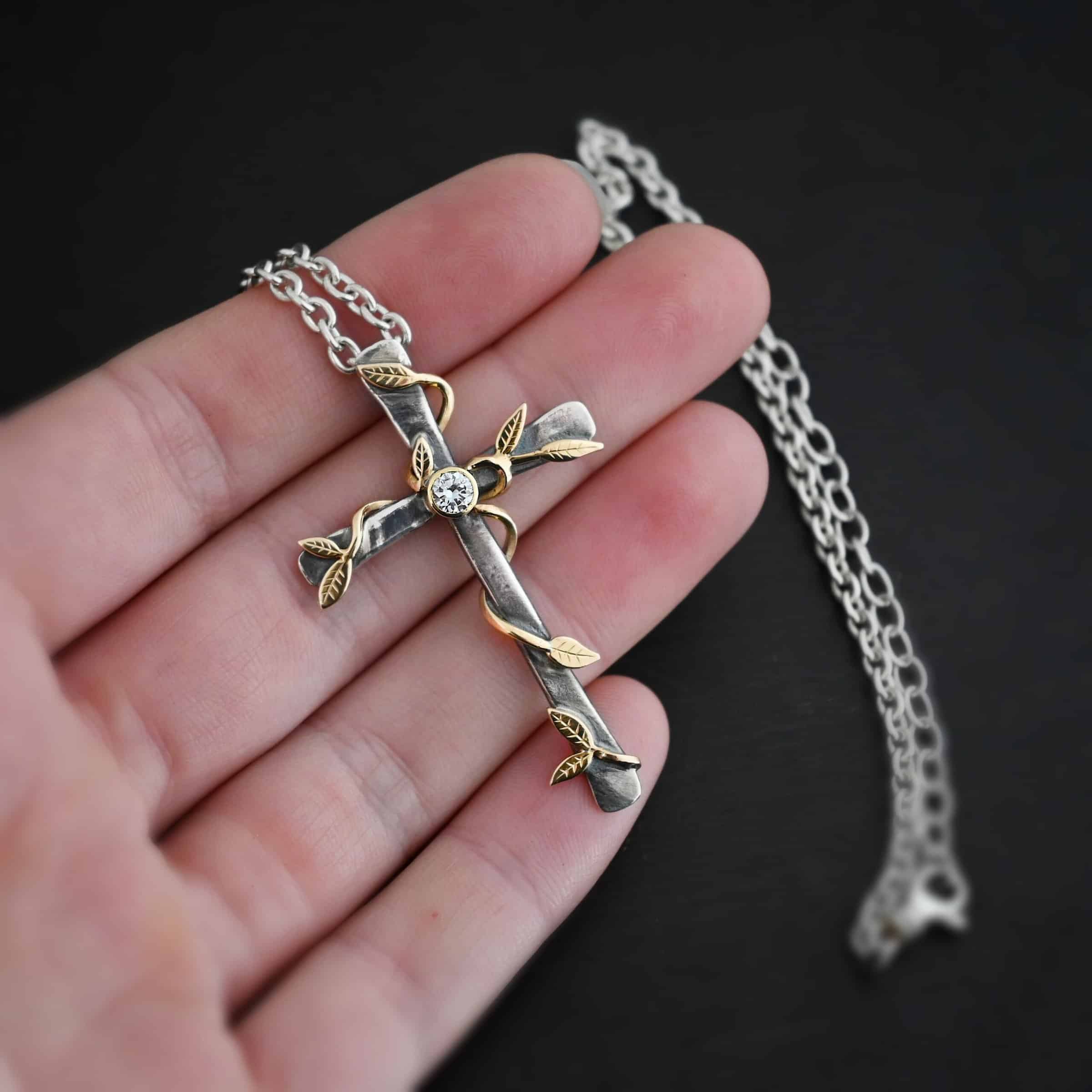 Photo of a custom "He Is Risen" Necklace. There is a large sized, smooth, oxidized, cross pendant. The center has a diamond set in gold. There are gold vines with leaves wrapped around the cross. Necklace is lying against the fingertips of a hand to demonstrate size.