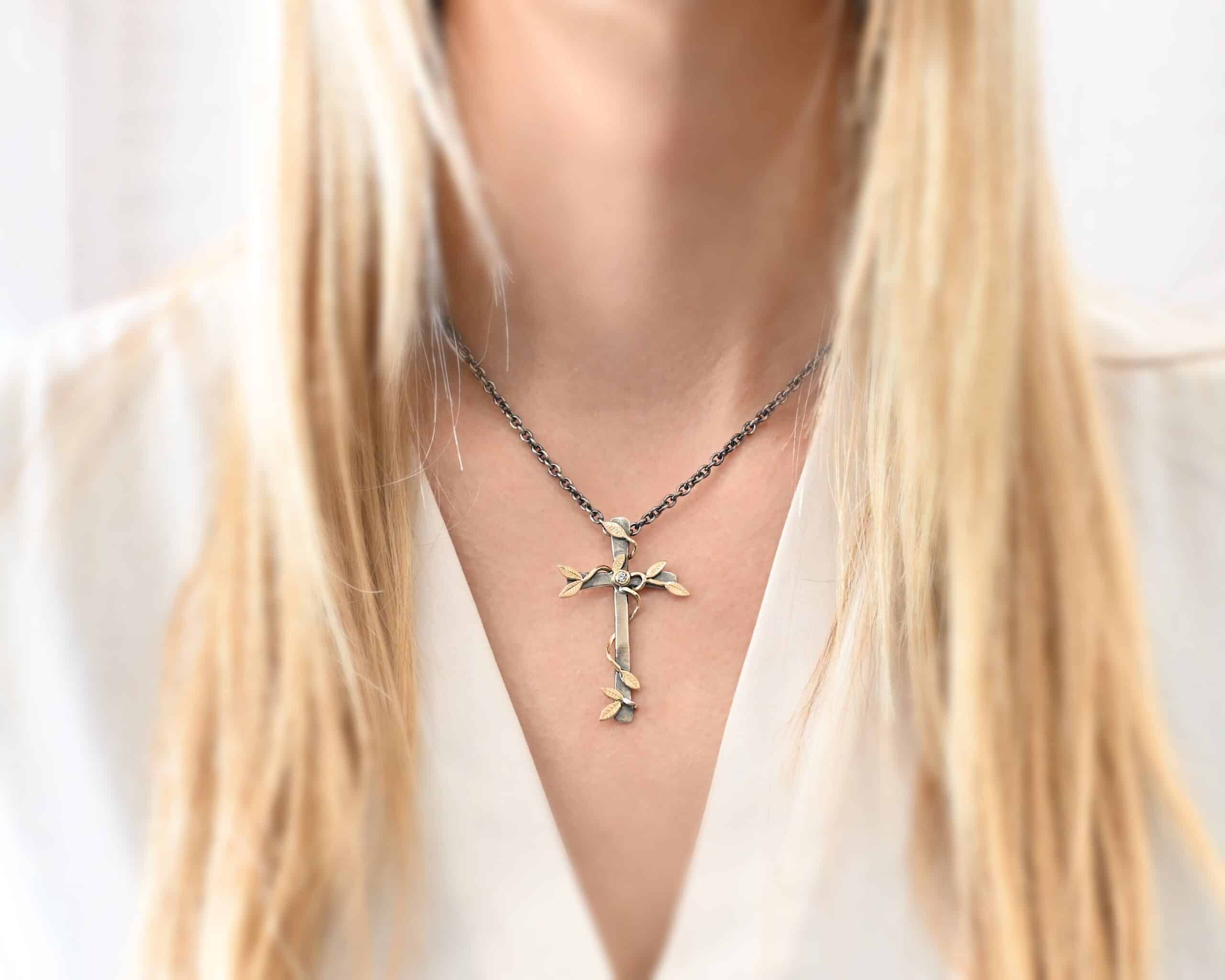 Photo of a custom "He Is Risen" Necklace. There is a large sized, smooth, oxidized, cross pendant. The center has a diamond set in gold. There are gold vines with leaves wrapped around the cross. Necklace is worn by a model to demonstrate size.
