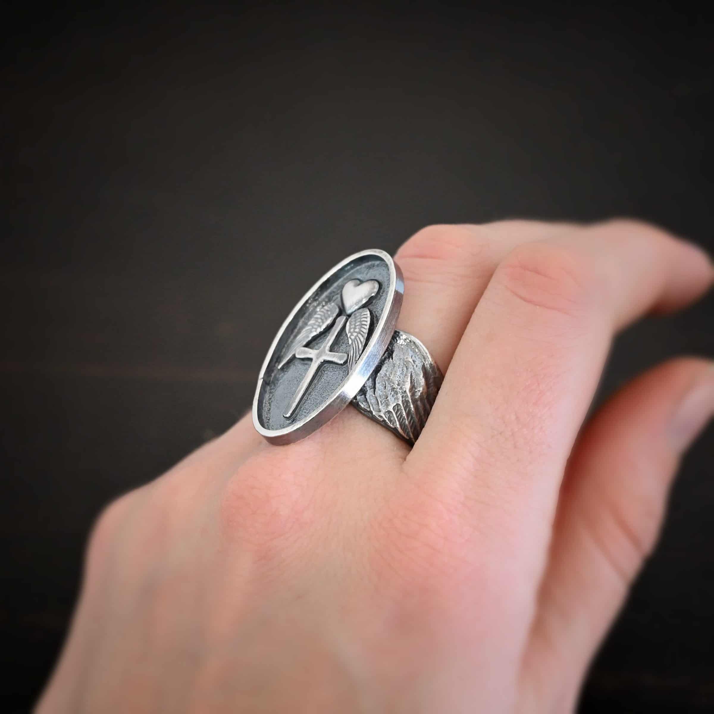 Photo of the "Sacred Heart" Ring. Made out of sterling silver. The ring has a large, oval shaped, thick piece of silver in the center of the ring. On the inside of the oval is a small heart at the top that sticks up so it is noticeable. There is an angel wing beneath the heart on each side, so two angel wings. Beneath the heart, on the bottom tip of it, is a cross that sticks up. The end of each angel wing touches the left and right side of the cross. The band of the ring is made out of curved sterling silver angel wings. Ring is worn by a model to demonstrate size.