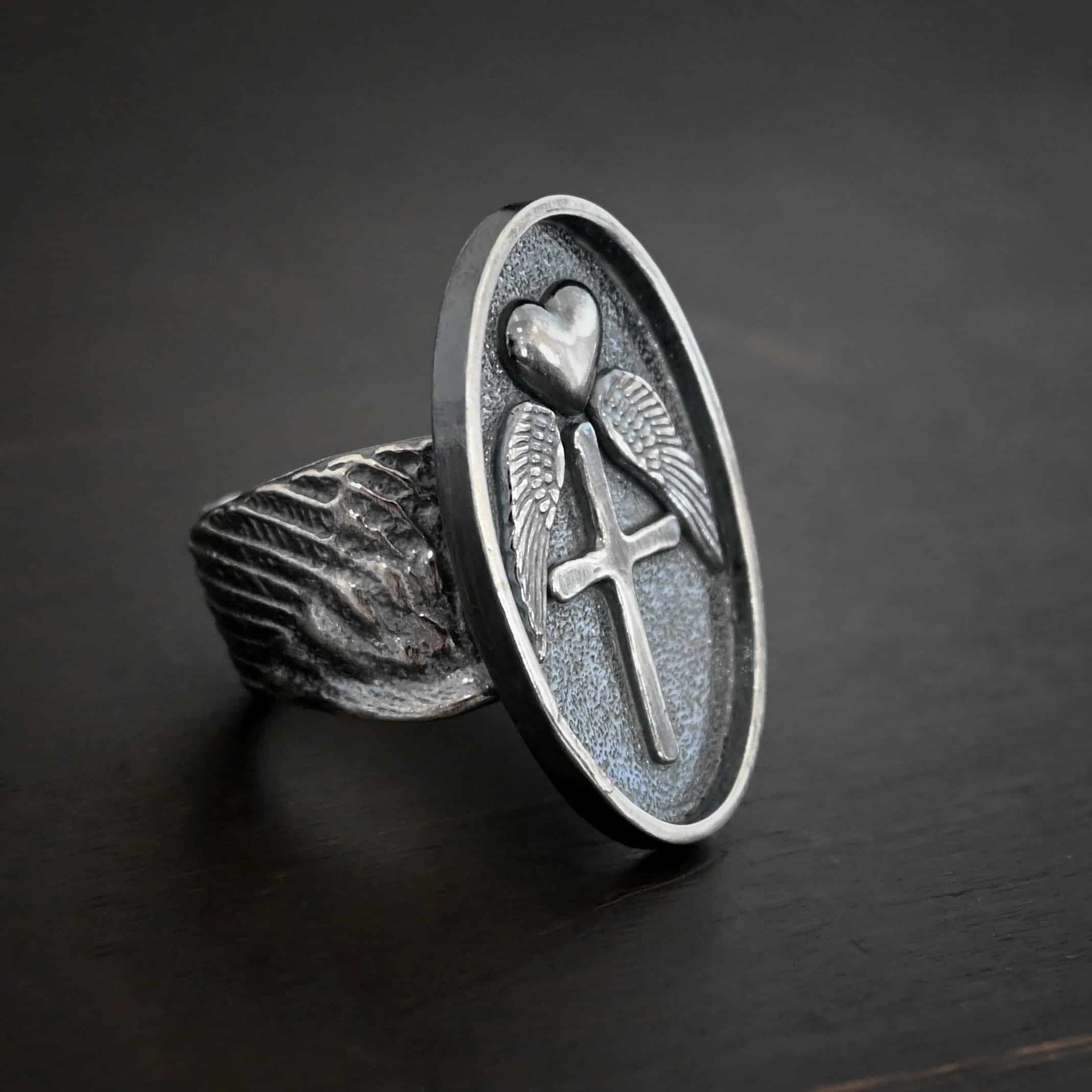 Photo of the "Sacred Heart" Ring. Made out of sterling silver. The ring has a large, oval shaped, thick piece of silver in the center of the ring. On the inside of the oval is a small heart at the top that sticks up so it is noticeable. There is an angel wing beneath the heart on each side, so two angel wings. Beneath the heart, on the bottom tip of it, is a cross that sticks up. The end of each angel wing touches the left and right side of the cross. The band of the ring is made out of curved sterling silver angel wings. Ring is sitting against a dark background.