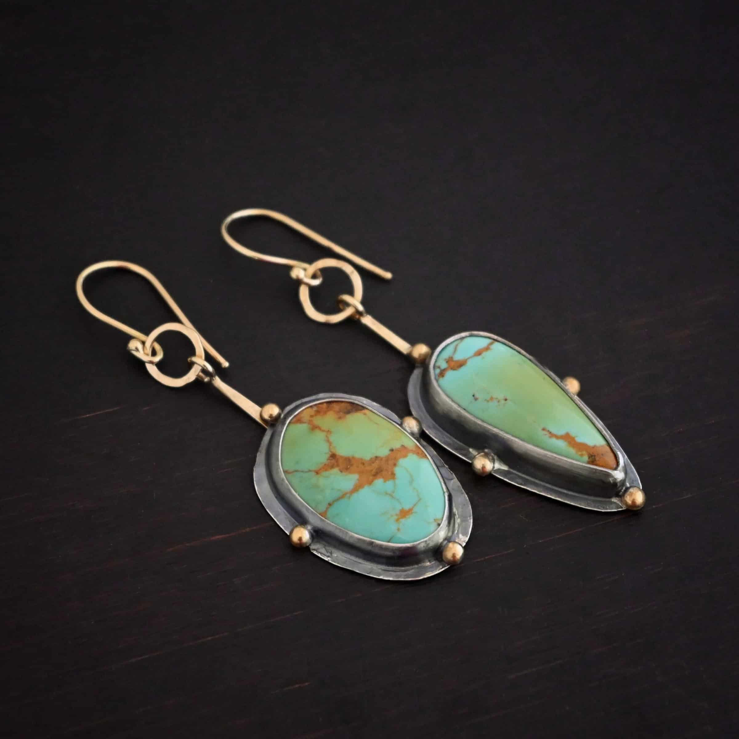 Gilded Azure Earrings Photo of the "Gilded Azure" Earrings. Made out of Sterling Silver & 18K Gold, & Kingman Turquoise. The earrings are gold french hook ear wires. There is a medium sized gold circle that is hammered flat going through the bottom circle of the ear wire. Hanging from the bottom of the gold circle is a small stick of gold with a loop at the top. The bottom of the stick is soldered to the back of the turquoise stone setting. One turquoise is an oval shape, and the other is a teardrop shape with the point facing down. Each stone has four gold balls going around it. One ball is on the top, one on the left side, one on the right, and one on the bottom. The turquoise are set in silver, and the silver around the stones is hammered flat, which is what the gold balls are on. Earrings are lying against a dark background.