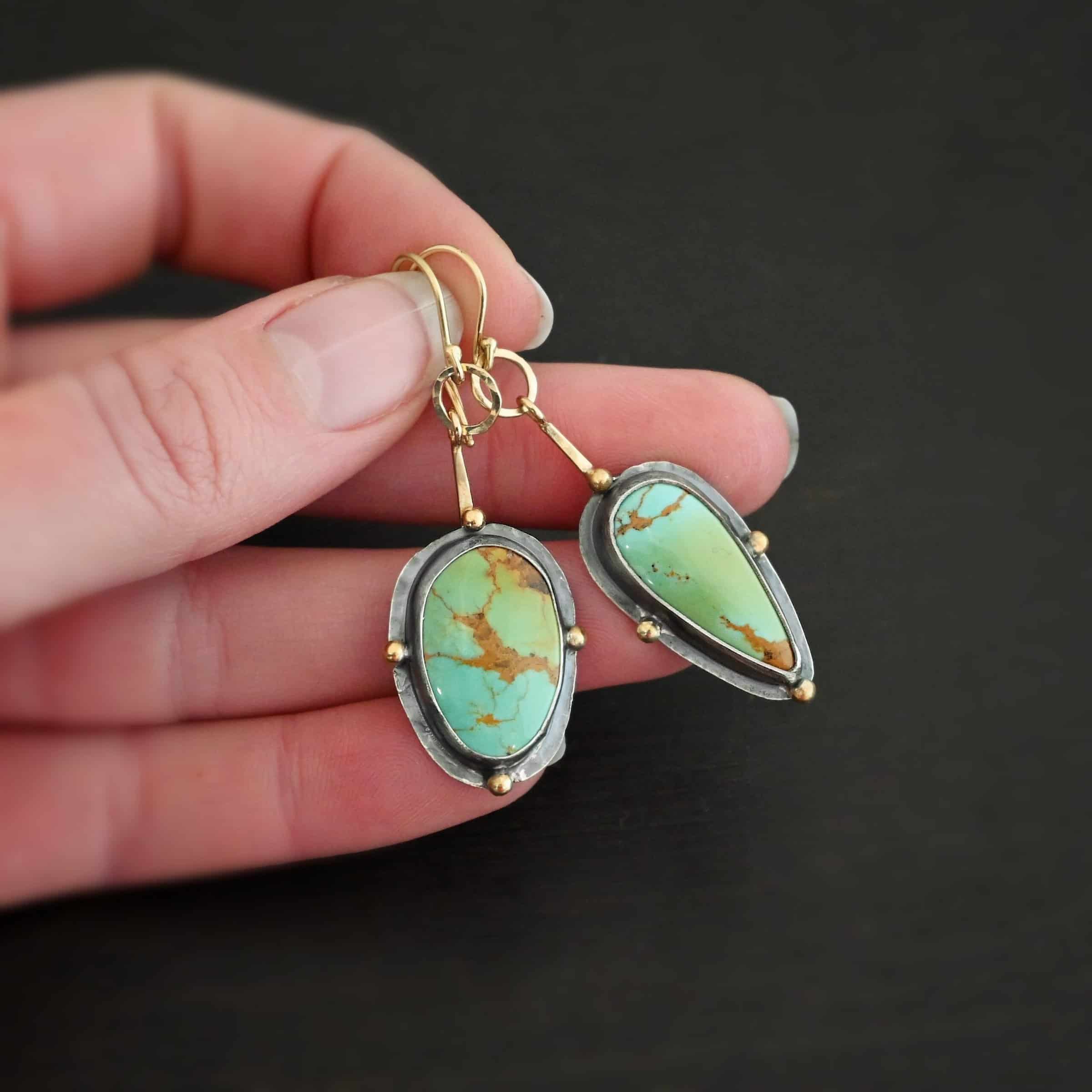 Gilded Azure Earrings Photo of the "Gilded Azure" Earrings. Made out of Sterling Silver & 18K Gold, & Kingman Turquoise. The earrings are gold french hook ear wires. There is a medium sized gold circle that is hammered flat going through the bottom circle of the ear wire. Hanging from the bottom of the gold circle is a small stick of gold with a loop at the top. The bottom of the stick is soldered to the back of the turquoise stone setting. One turquoise is an oval shape, and the other is a teardrop shape with the point facing down. Each stone has four gold balls going around it. One ball is on the top, one on the left side, one on the right, and one on the bottom. The turquoise are set in silver, and the silver around the stones is hammered flat, which is what the gold balls are on. Earrings are held in the fingertips of a hand to demonstrate size.