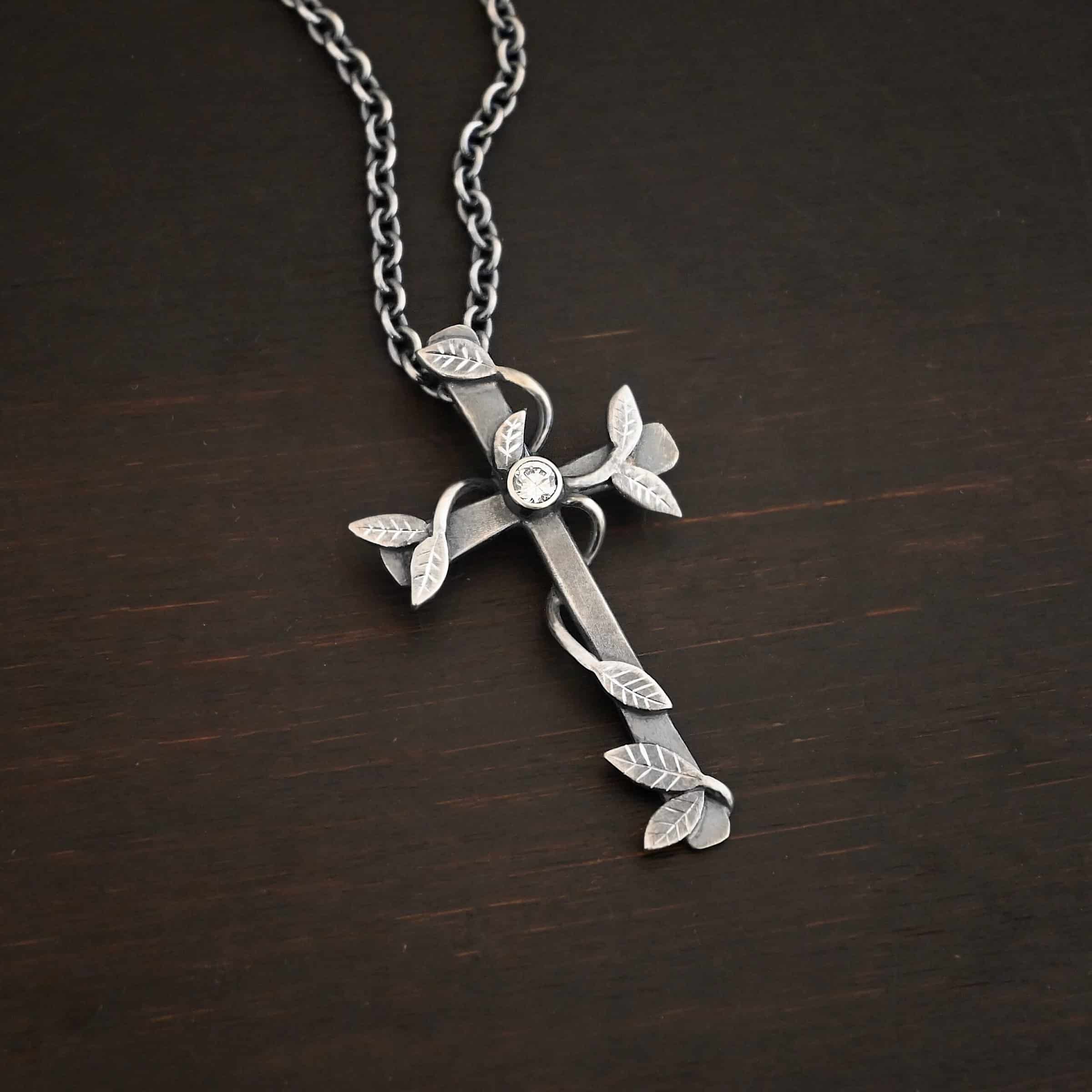 Photo of the "He Is Risen" Necklace. Necklace is made out of sterling silver and diamond. The cross is made out of sterling silver that is smooth and oxidized. There are silver vines wrapped around the entire cross with small silver leaves on the ends of the vines. There is a diamond in the center of the cross that is set in silver. There is a loop soldered to the back of the necklace that the chain of the necklace goes through. Necklace is lying against a dark background.