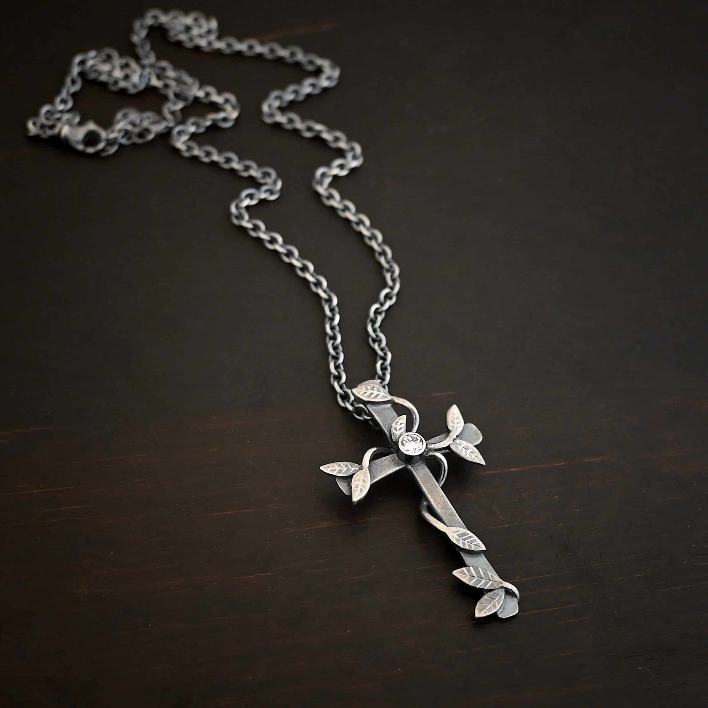 Photo of the "He Is Risen" Necklace. Necklace is made out of sterling silver and diamond. The cross is made out of sterling silver that is smooth and oxidized. There are silver vines wrapped around the entire cross with small silver leaves on the ends of the vines. There is a diamond in the center of the cross that is set in silver. There is a loop soldered to the back of the necklace that the chain of the necklace goes through. Necklace is lying against a dark background.