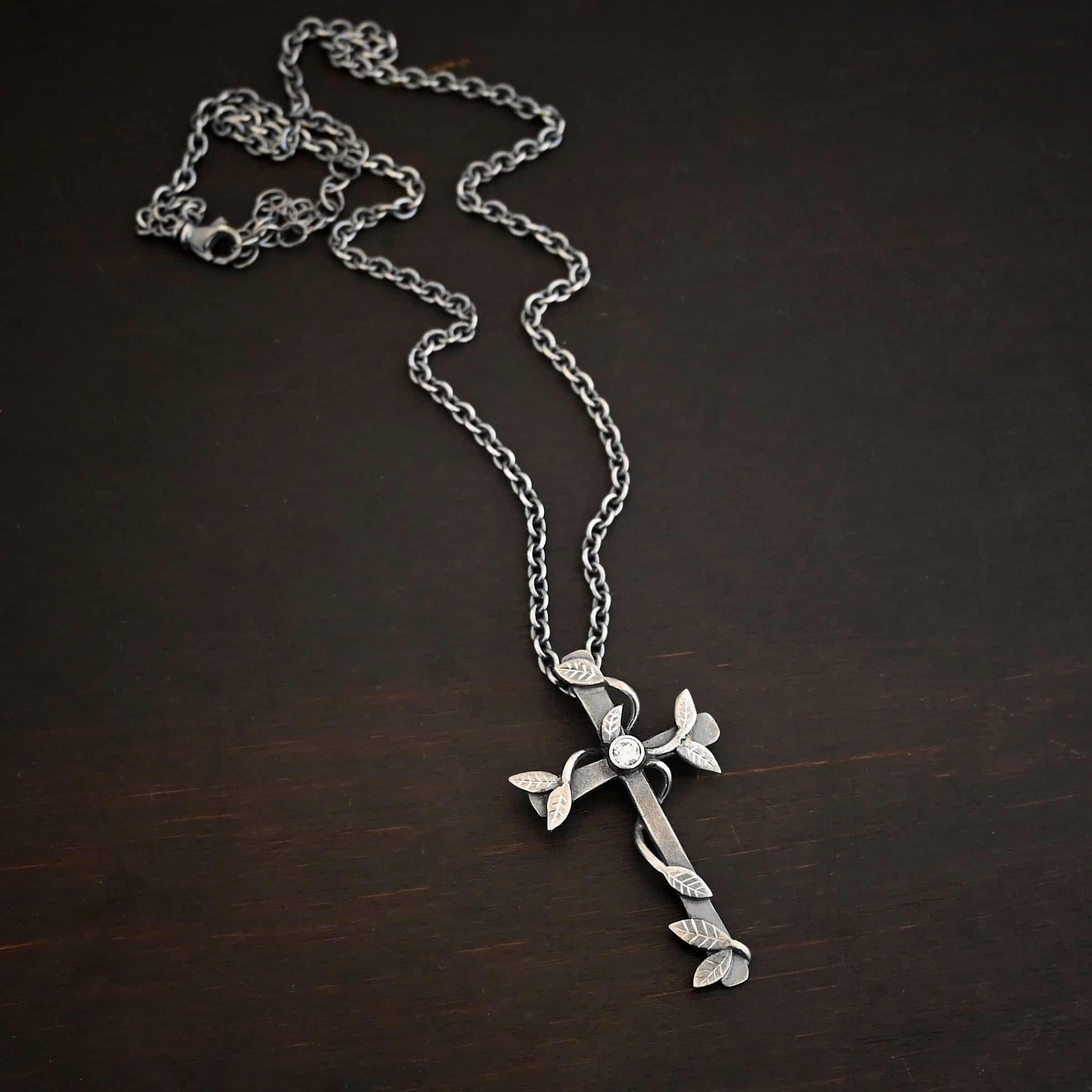 Photo of the "He Is Risen" Necklace. Necklace is made out of sterling silver and diamond. The cross is made out of sterling silver that is smooth and oxidized. There are silver vines wrapped around the entire cross with small silver leaves on the ends of the vines. There is a diamond in the center of the cross that is set in silver. There is a loop soldered to the back of the necklace that the chain of the necklace goes through. Necklace is lying against a dark background.