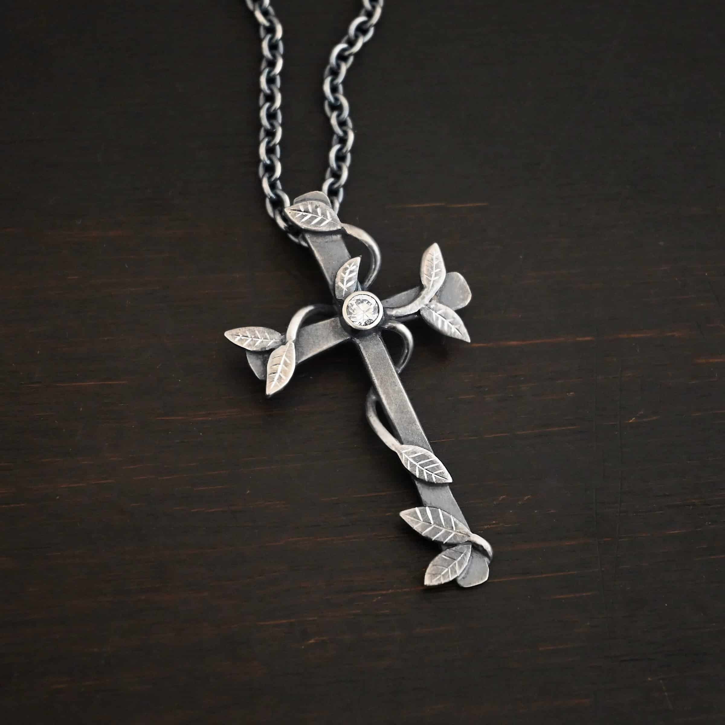 Photo of the "He Is Risen" Necklace. Necklace is made out of sterling silver and diamond. The cross is made out of sterling silver that is smooth and oxidized. There are silver vines wrapped around the entire cross with small silver leaves on the ends of the vines. There is a diamond in the center of the cross that is set in silver. There is a loop soldered to the back of the necklace that the chain of the necklace goes through. Necklace is lying against a dark background.
