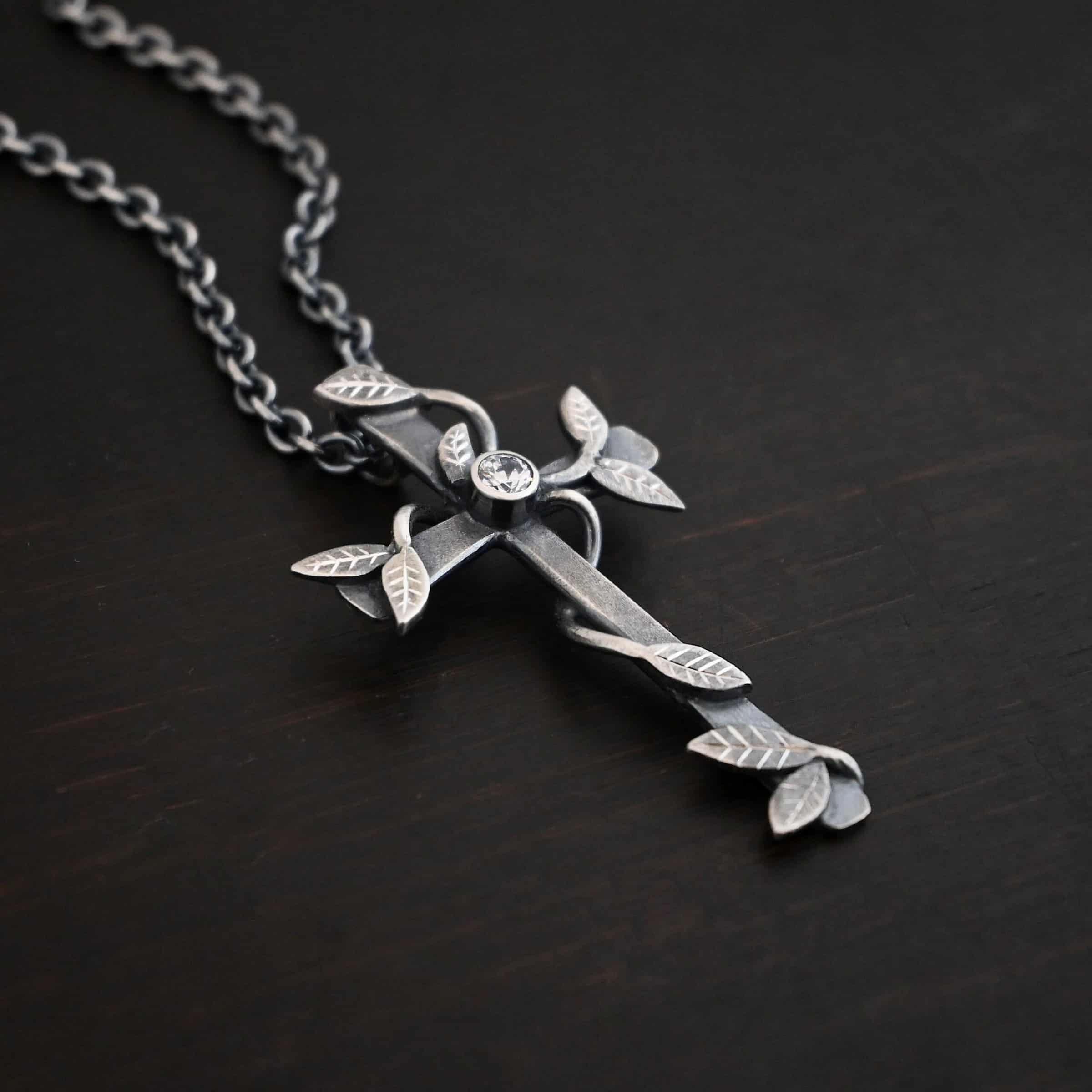 Photo of the "He Is Risen" Necklace. Necklace is made out of sterling silver and diamond. The cross is made out of sterling silver that is smooth and oxidized. There are silver vines wrapped around the entire cross with small silver leaves on the ends of the vines. There is a diamond in the center of the cross that is set in silver. There is a loop soldered to the back of the necklace that the chain of the necklace goes through. Necklace is lying against a dark background.