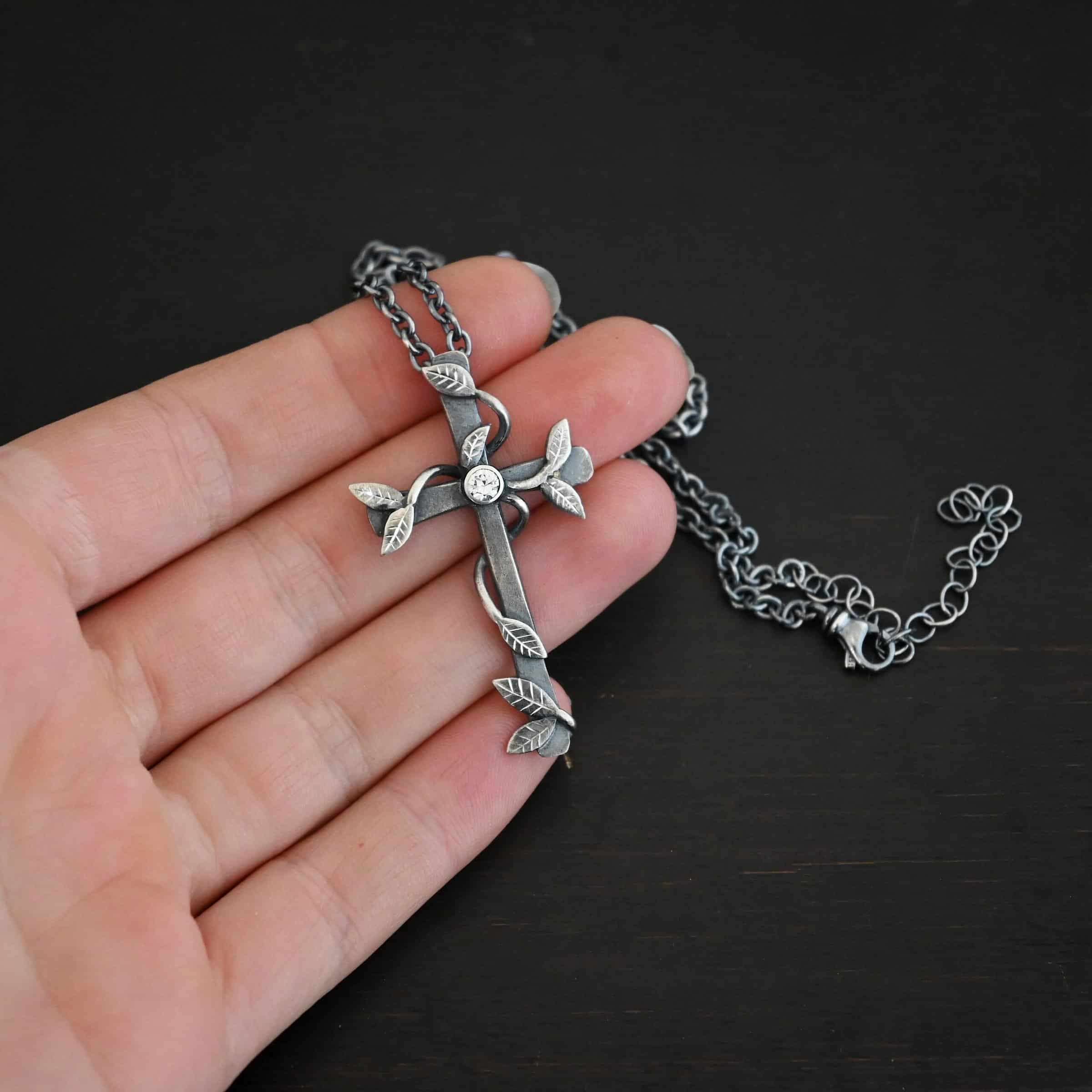 Photo of the "He Is Risen" Necklace. Necklace is made out of sterling silver and diamond. The cross is made out of sterling silver that is smooth and oxidized. There are silver vines wrapped around the entire cross with small silver leaves on the ends of the vines. There is a diamond in the center of the cross that is set in silver. There is a loop soldered to the back of the necklace that the chain of the necklace goes through. Necklace is lying against the fingertips of a hand to demonstrate size.