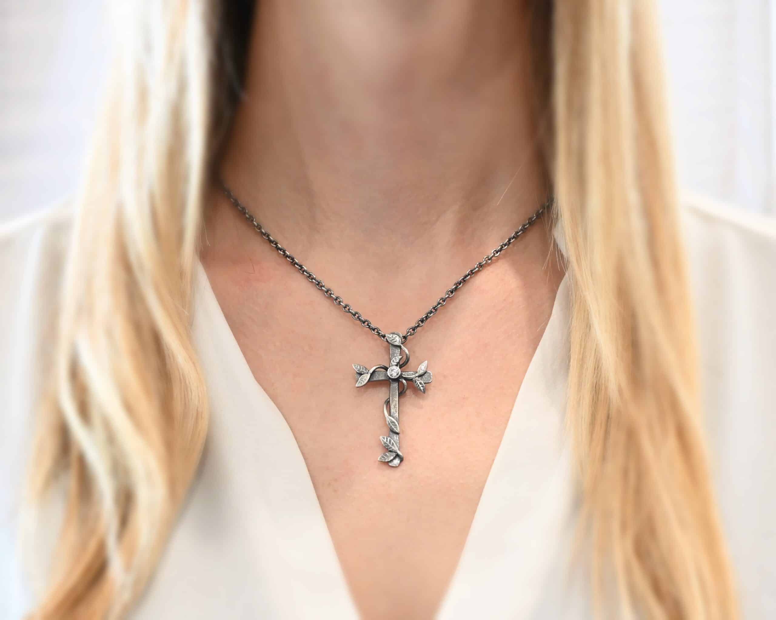 Photo of the "He Is Risen" Necklace shown on a model. Necklace is made out of sterling silver and diamond. The cross is made out of sterling silver that is smooth and oxidized. There are silver vines wrapped around the entire cross with small silver leaves on the ends of the vines. There is a diamond in the center of the cross that is set in silver. There is a loop soldered to the back of the necklace that the chain of the necklace goes through. Model is wearing it to demonstrate size.