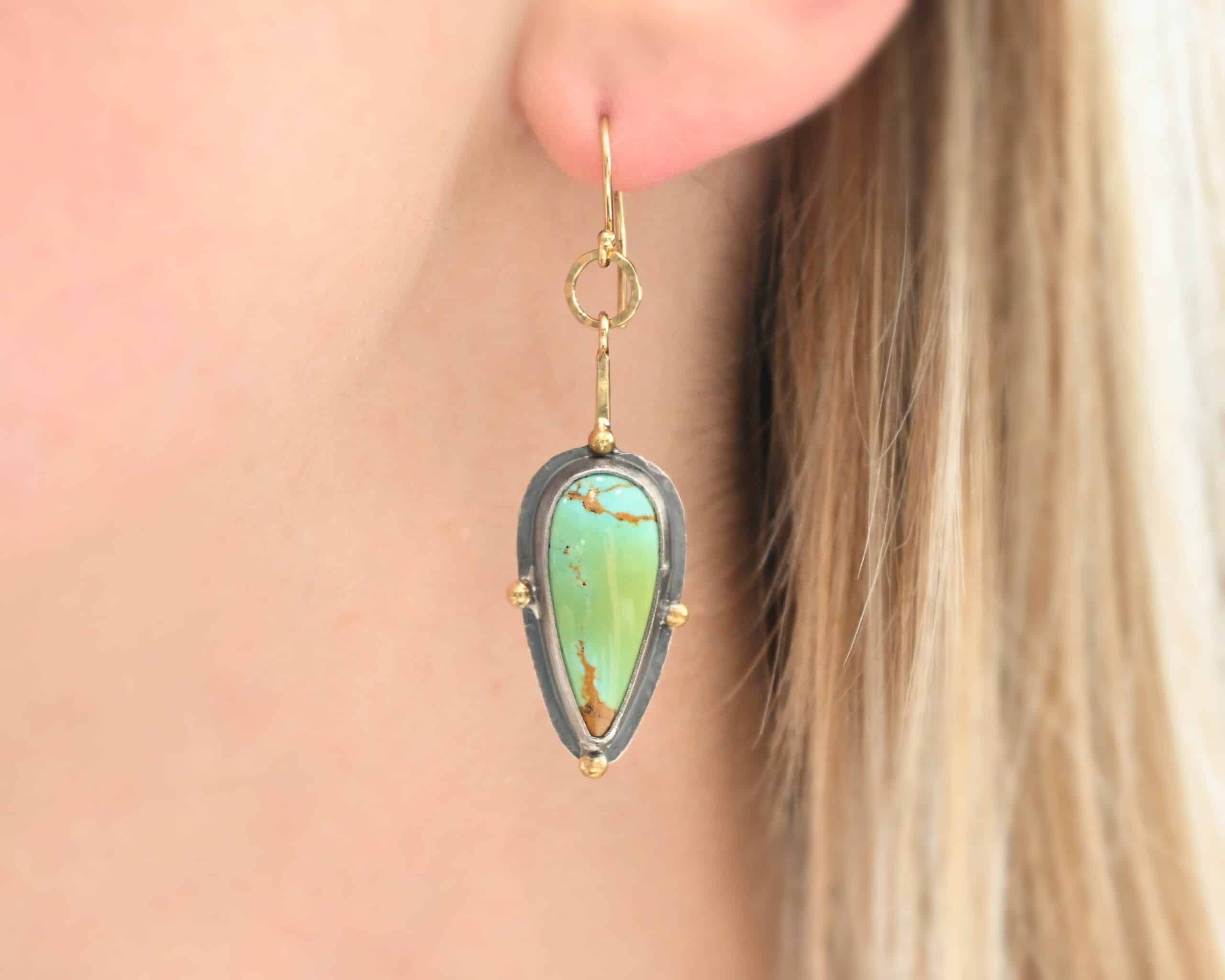 Gilded Azure Earrings Photo of the "Gilded Azure" Earring as worn on a model. Made out of Sterling Silver & 18K Gold, & Kingman Turquoise. The earring is on a gold french hook ear wire. There is a medium sized gold circle that is hammered flat going through the bottom circle of the ear wire. Hanging from the bottom of the gold circle is a small stick of gold with a loop at the top. The bottom of the stick is soldered to the back of the turquoise stone setting. The turquoise is a teardrop shape with the point facing down. The stone has four gold balls going around it. One ball is on the top, one on the left side, one on the right, and one on the bottom. The turquoise is set in silver, and the silver around the stones is hammered flat, which is what the gold balls are on.