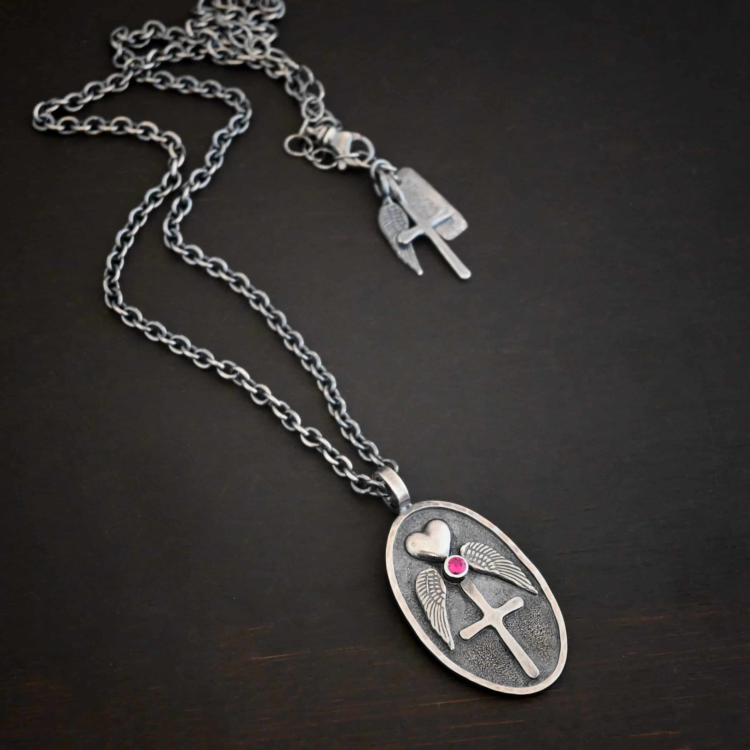 My Forever Angel Necklace Photo of the "My Forever Angel" Necklace. Made out of sterling silver. The necklace has a large, oval shaped, thick piece of silver for the pendant. On the inside of the oval is a small heart at the top that sticks up so it is noticeable. There is a small ruby on the bottom tip of the heart. There is an angel wing beneath the heart on each side, surrounding the ruby. Each wing faces outwards. Beneath the diamond is a cross that is soldered. The end of each angel wing touches the left and right side of the cross. There is a thick silver loop soldered to the top. The chain of the necklace goes through the loop. Necklace is lying against a dark background.