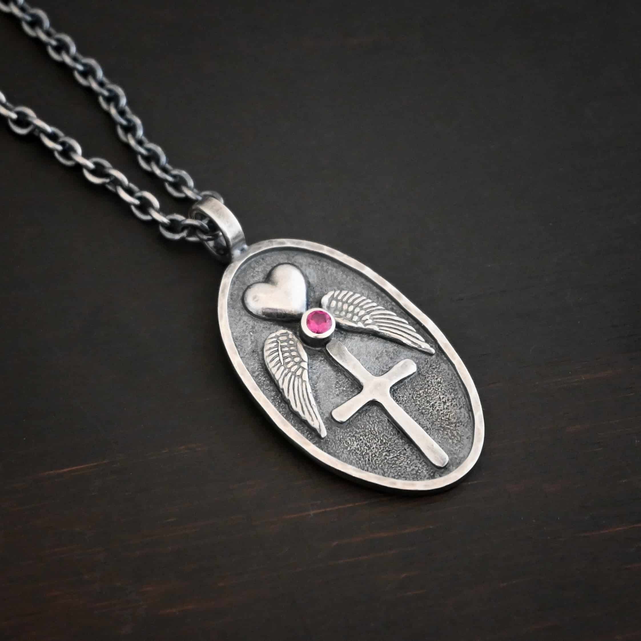 My Forever Angel Necklace Photo of the "My Forever Angel" Necklace. Made out of sterling silver. The necklace has a large, oval shaped, thick piece of silver for the pendant. On the inside of the oval is a small heart at the top that sticks up so it is noticeable. There is a small ruby on the bottom tip of the heart. There is an angel wing beneath the heart on each side, surrounding the ruby. Each wing faces outwards. Beneath the diamond is a cross that is soldered. The end of each angel wing touches the left and right side of the cross. There is a thick silver loop soldered to the top. The chain of the necklace goes through the loop. Necklace is lying against a dark background.