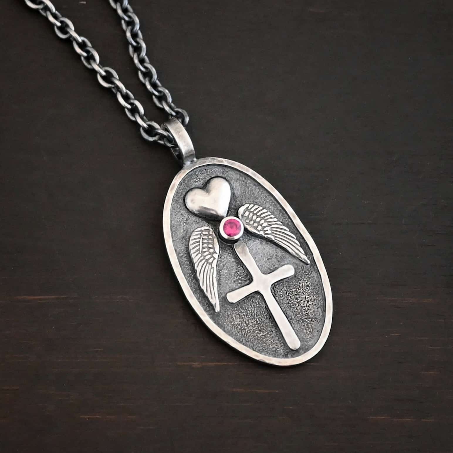 My Forever Angel Necklace Photo of the "My Forever Angel" Necklace. Made out of sterling silver. The necklace has a large, oval shaped, thick piece of silver for the pendant. On the inside of the oval is a small heart at the top that sticks up so it is noticeable. There is a small ruby on the bottom tip of the heart. There is an angel wing beneath the heart on each side, surrounding the ruby. Each wing faces outwards. Beneath the diamond is a cross that is soldered. The end of each angel wing touches the left and right side of the cross. There is a thick silver loop soldered to the top. The chain of the necklace goes through the loop. Necklace is lying against a dark background.