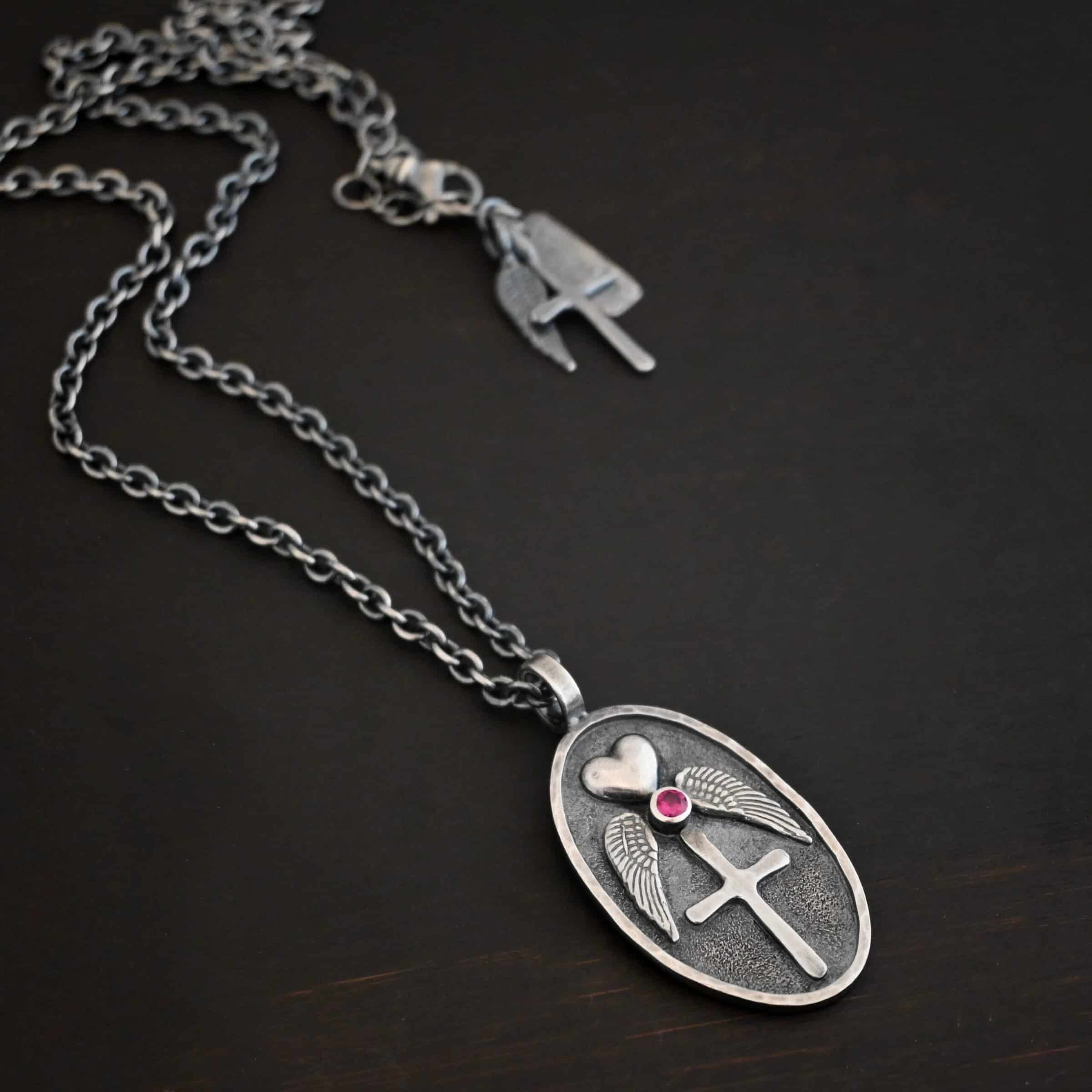 My Forever Angel Necklace Photo of the "My Forever Angel" Necklace. Made out of sterling silver. The necklace has a large, oval shaped, thick piece of silver for the pendant. On the inside of the oval is a small heart at the top that sticks up so it is noticeable. There is a small ruby on the bottom tip of the heart. There is an angel wing beneath the heart on each side, surrounding the ruby. Each wing faces outwards. Beneath the diamond is a cross that is soldered. The end of each angel wing touches the left and right side of the cross. There is a thick silver loop soldered to the top. The chain of the necklace goes through the loop. Necklace is lying against a dark background.