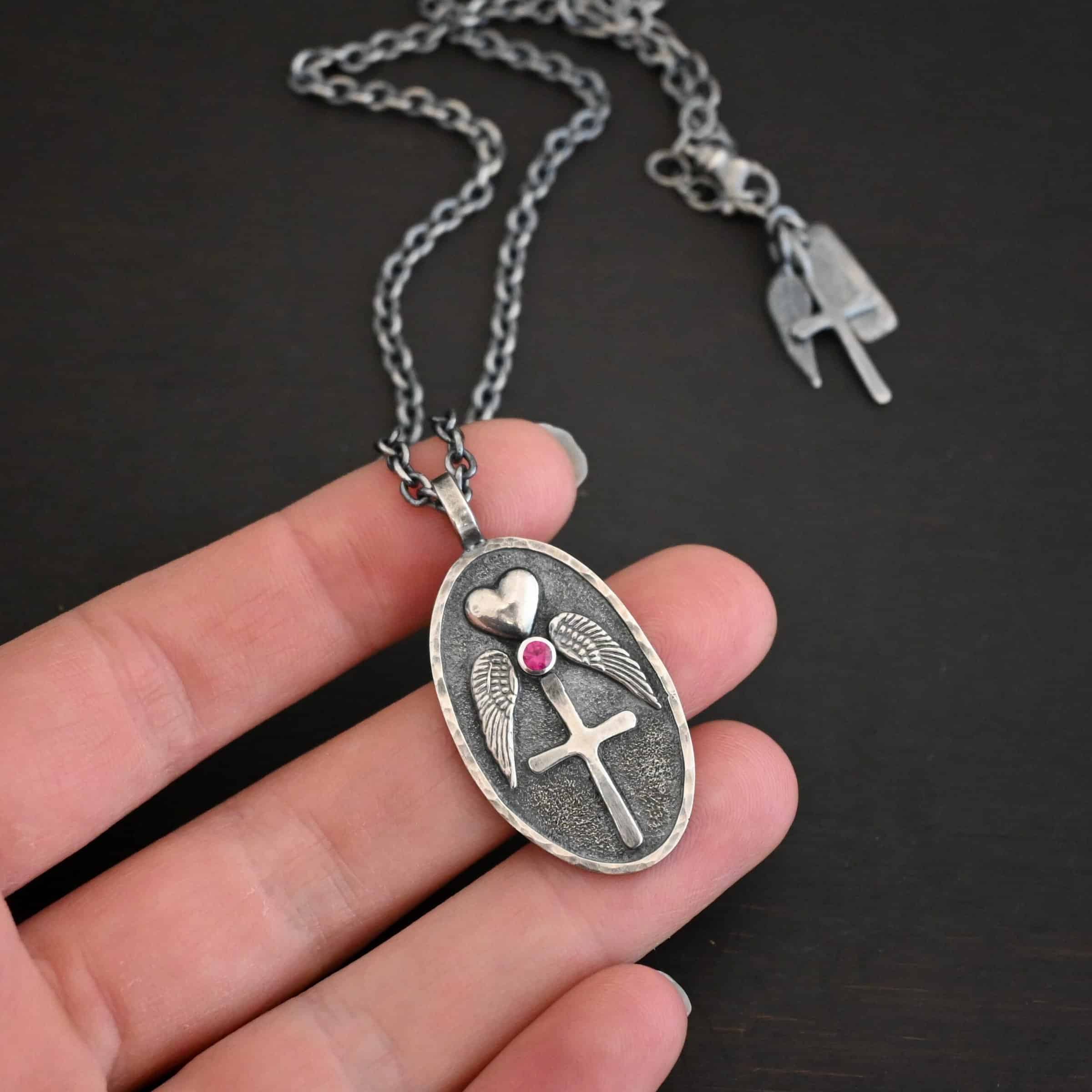 My Forever Angel Necklace Photo of the "My Forever Angel" Necklace. Made out of sterling silver. The necklace has a large, oval shaped, thick piece of silver for the pendant. On the inside of the oval is a small heart at the top that sticks up so it is noticeable. There is a small ruby on the bottom tip of the heart. There is an angel wing beneath the heart on each side, surrounding the ruby. Each wing faces outwards. Beneath the diamond is a cross that is soldered. The end of each angel wing touches the left and right side of the cross. There is a thick silver loop soldered to the top. The chain of the necklace goes through the loop. Necklace is lying against the fingertips of a hand.