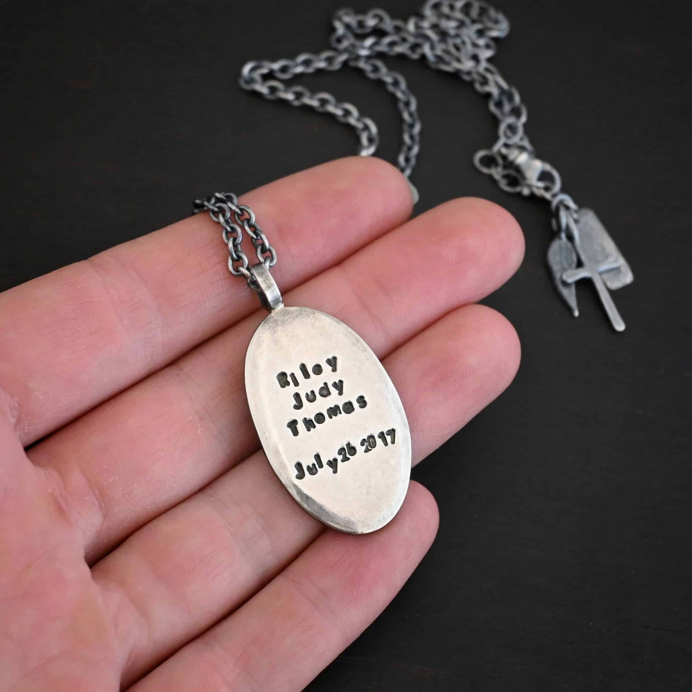 My Forever Angel Necklace Photo of the back of the "My Forever Angel" Necklace. Made out of sterling silver. The necklace has a large, oval shaped, thick piece of silver for the pendant. The back of the pendant is smooth silver. It has the name "Riley Judy Thomas" stamped into it, and the date "July 26 2017" stamped into it as well. Both the name and date are darkened to stand out. Necklace is lying against the fingertips of a hand to demonstrate size.