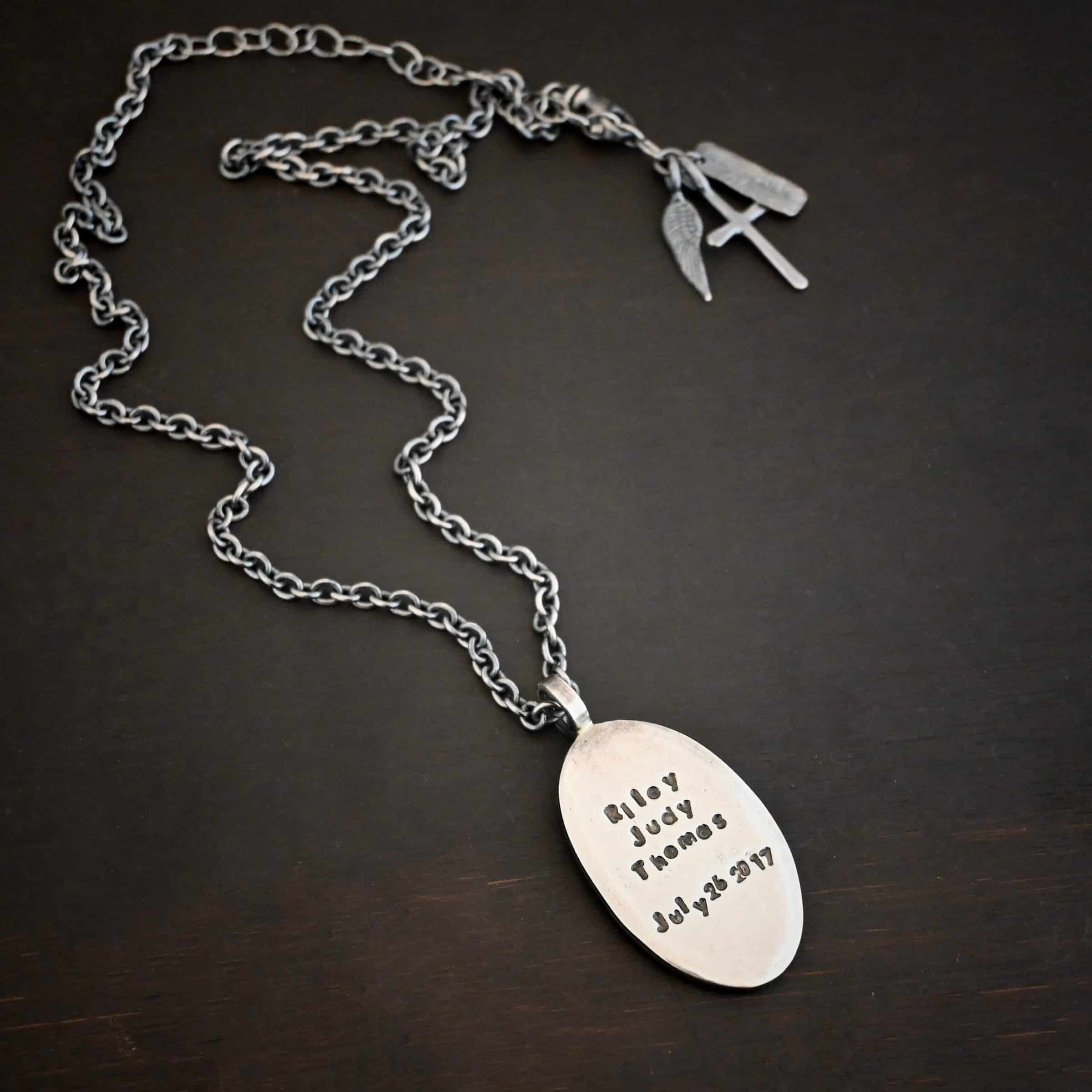My Forever Angel Necklace Photo of the back of the "My Forever Angel" Necklace. Made out of sterling silver. The necklace has a large, oval shaped, thick piece of silver for the pendant. The back of the pendant is smooth silver. It has the name "Riley Judy Thomas" stamped into it, and the date "July 26 2017" stamped into it as well. Both the name and date are darkened to stand out. Necklace is lying against a dark background.