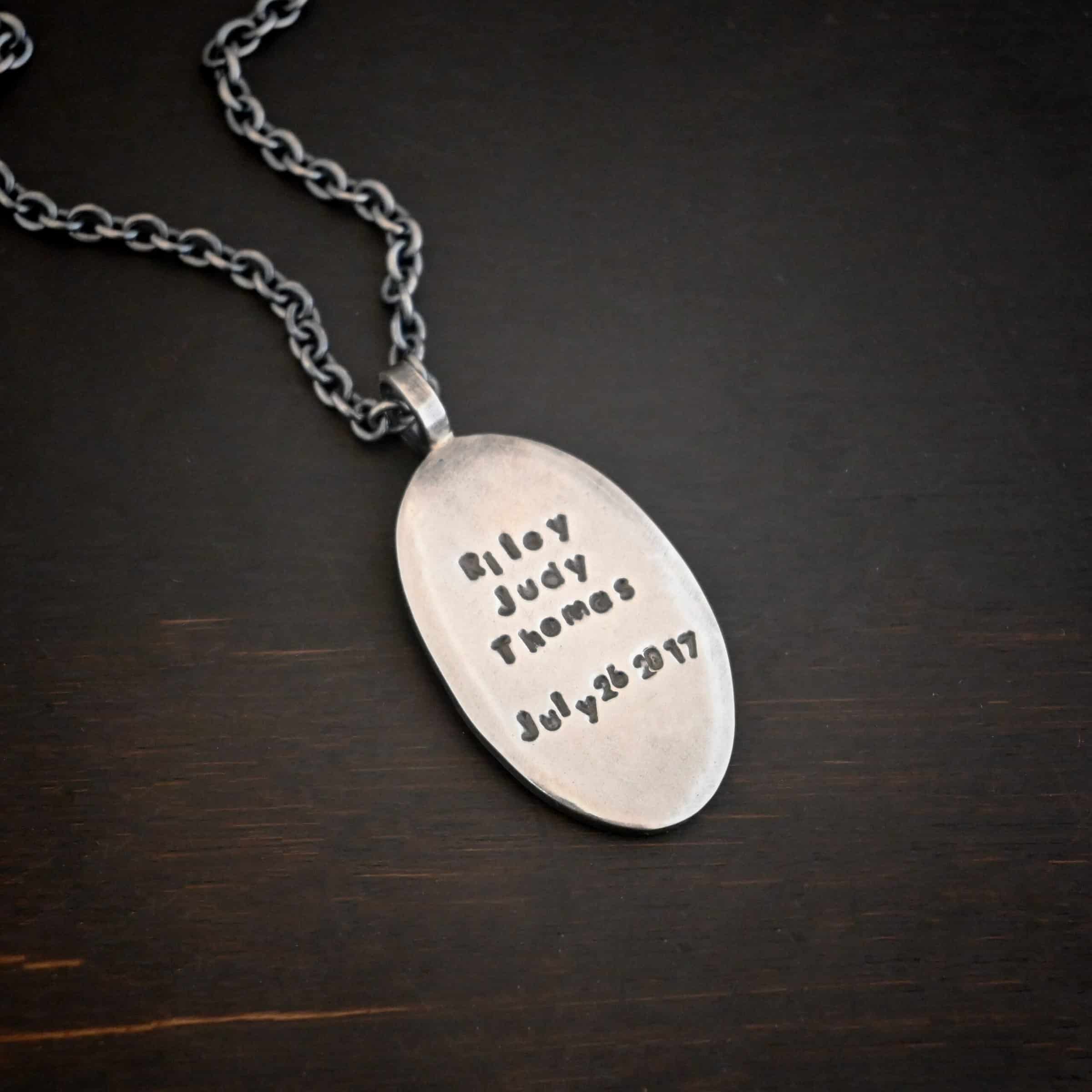 My Forever Angel Necklace Photo of the back of the "My Forever Angel" Necklace. Made out of sterling silver. The necklace has a large, oval shaped, thick piece of silver for the pendant. The back of the pendant is smooth silver. It has the name "Riley Judy Thomas" stamped into it, and the date "July 26 2017" stamped into it as well. Both the name and date are darkened to stand out. Necklace is lying against a dark background.