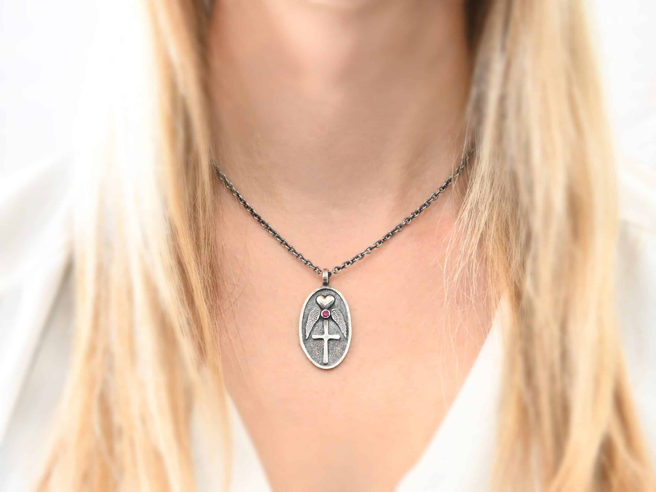 My Angel With Me Memorial Necklace Photo of the "My Angel With Me" Necklace. Made out of sterling silver. The necklace has a large, oval shaped, thick piece of silver for the pendant. On the inside of the oval is a small heart at the top that sticks up so it is noticeable. There is a small ruby on the bottom tip of the heart. There is an angel wing beneath the heart on each side, surrounding the ruby. Each wing faces outwards. Beneath the diamond is a cross that is soldered. The end of each angel wing touches the left and right side of the cross. There is a thick silver loop soldered to the top. The chain of the necklace goes through the loop. Necklace is worn by a model to demonstrate size.