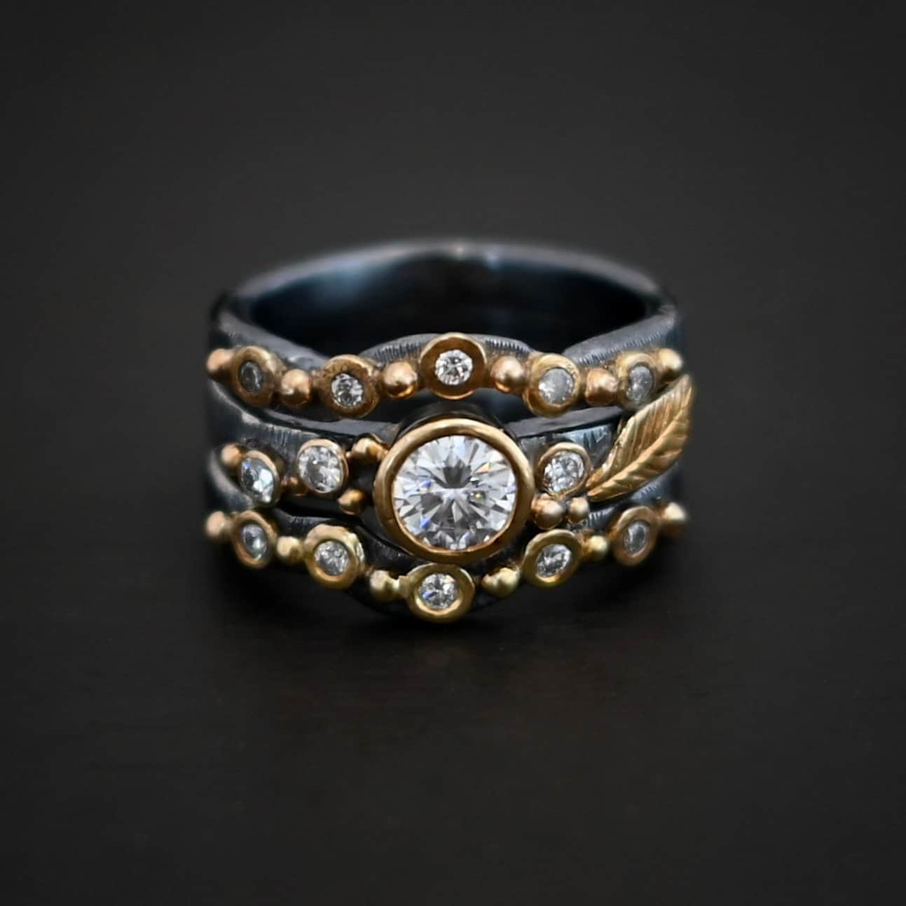 Photo of the "Forever Held" Ring. Ring has a 1.5 carat moissanite in the center, with two diamonds on the left side of it and one on the right. There is gold ball detail and a gold leaf on the band as well. Ring has a top and bottom band that have a dip in the center to fit around the center stone. These bands have multiple diamonds around them with balls of gold that separate each diamond. Ring is sitting against a dark background.