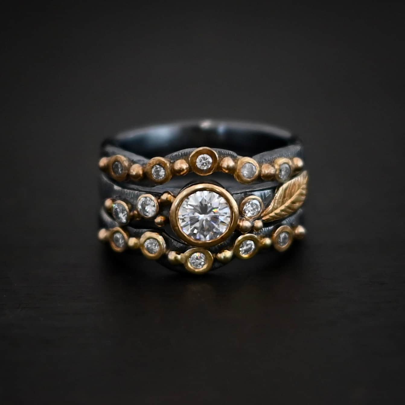 Photo of the "Forever Held" Ring. Ring has a 1.5 carat moissanite in the center, with two diamonds on the left side of it and one on the right. There is gold ball detail and a gold leaf on the band as well. Ring has a top and bottom band that have a dip in the center to fit around the center stone. These bands have multiple diamonds around them with balls of gold that separate each diamond. Ring is sitting against a dark background.